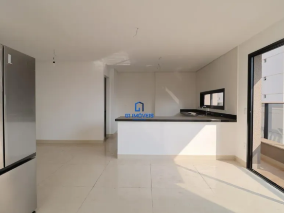 A imagem mostra um espaço interno que parece ser a cozinha e sala de estar de um apartamento ou casa recém-construída ou reformada. O ambiente é caracterizado por cores neutras, com paredes brancas e piso claro, possivelmente de cerâmica ou porcelanato, que refletem a luz e ampliam a sensação de espaço. À esquerda, destaca-se uma geladeira de aço inoxidável moderna e grande, com múltiplas portas, o que sugere um eletrodoméstico de alta qualidade. A geladeira parece ser um dos poucos objetos presentes, indicando que o espaço está pronto para ser mobiliado. Ao fundo, há uma bancada que separa a cozinha da sala de estar. A parte inferior da bancada é branca, enquanto o tampo é preto, proporcionando um contraste elegante e contemporâneo. Na bancada, observa-se uma pia e uma torneira, indicando que essa área é destinada à preparação de alimentos. Uma pequena janela acima da pia oferece iluminação natural. A porta de vidro deslizante à direita leva a uma varanda, com vista para outros edifícios, sugerindo que o imóvel está localizado em uma área urbana densa. A moldura preta da porta de vidro adiciona um toque moderno e sofisticado ao espaço. O espaço parece ser amplo e bem iluminado, com um design minimalista e funcional. A ausência de móveis e decoração permite que o futuro morador personalize o ambiente de acordo com suas preferências. A imagem transmite uma sensação de limpeza, organização e modernidade, tornando o imóvel atraente para potenciais compradores ou inquilinos.