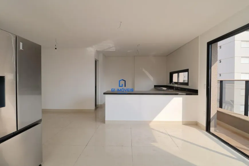 A imagem mostra um espaço interno que parece ser a cozinha e sala de estar de um apartamento ou casa recém-construída ou reformada. O ambiente é caracterizado por cores neutras, com paredes brancas e piso claro, possivelmente de cerâmica ou porcelanato, que refletem a luz e ampliam a sensação de espaço. À esquerda, destaca-se uma geladeira de aço inoxidável moderna e grande, com múltiplas portas, o que sugere um eletrodoméstico de alta qualidade. A geladeira parece ser um dos poucos objetos presentes, indicando que o espaço está pronto para ser mobiliado. Ao fundo, há uma bancada que separa a cozinha da sala de estar. A parte inferior da bancada é branca, enquanto o tampo é preto, proporcionando um contraste elegante e contemporâneo. Na bancada, observa-se uma pia e uma torneira, indicando que essa área é destinada à preparação de alimentos. Uma pequena janela acima da pia oferece iluminação natural. A porta de vidro deslizante à direita leva a uma varanda, com vista para outros edifícios, sugerindo que o imóvel está localizado em uma área urbana densa. A moldura preta da porta de vidro adiciona um toque moderno e sofisticado ao espaço. O espaço parece ser amplo e bem iluminado, com um design minimalista e funcional. A ausência de móveis e decoração permite que o futuro morador personalize o ambiente de acordo com suas preferências. A imagem transmite uma sensação de limpeza, organização e modernidade, tornando o imóvel atraente para potenciais compradores ou inquilinos.