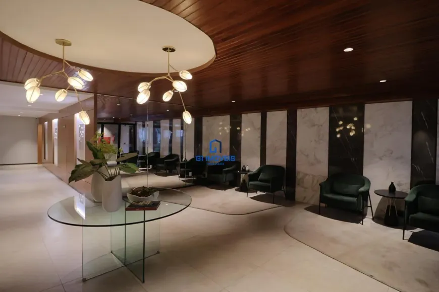 Piso: Material: Analisando a textura e o brilho, é provável que o piso seja feito de porcelanato polido de grande formato, imitando mármore. A ausência de porosidade visível sugere um acabamento impecável e fácil de limpar. Cor e Padrão: O bege suave com nuances creme apresenta variações sutis, imitando os padrões naturais do mármore. É possível observar veios delicados em tons mais claros, quebrando a monotonia e adicionando profundidade visual. Juntas: As juntas são tão finas que quase desaparecem, criando um efeito de continuidade e amplitude. O rejunte utilizado provavelmente é da mesma cor do piso, reforçando a uniformidade. Mesa Central: Tampo: O vidro temperado do tampo da mesa é cristalino e translúcido, permitindo que a luz o atravesse sem distorções. As bordas são polidas e arredondadas, garantindo segurança e um toque suave. Base: A base em forma de "X" é feita de vidro mais espesso, o que confere estabilidade e resistência à mesa. As peças de vidro são unidas por meio de encaixes precisos e discretos, sem parafusos ou colas visíveis. Detalhes: É possível que a mesa possua pequenos niveladores de altura nos pés, garantindo que ela fique perfeitamente estável, mesmo em pisos ligeiramente irregulares. Arranjo de Mesa: Vasos: Os vasos brancos são feitos de cerâmica de alta qualidade, com acabamento fosco ou acetinado. O design é minimalista, com linhas simples e elegantes. Plantas: As plantas parecem ser de espécies variadas, com diferentes tons de verde e texturas. Algumas podem ser folhagens tropicais, como filodendros ou zamioculcas, conhecidas por sua resistência e beleza. Livro: O livro aberto sobre a mesa tem uma capa escura, possivelmente em couro ou tecido aveludado. As páginas internas parecem ser de papel de alta gramatura, com impressão de qualidade. Composição: A disposição dos vasos, plantas e livro é cuidadosamente pensada para criar um ponto focal interessante e equilibrado. A altura dos elementos varia, criando uma dinâmica visual agradável. Iluminação: Rebaixo: O rebaixo no teto possui bordas arredondadas e um acabamento impecável. A iluminação embutida é feita com lâmpadas LED de alta eficiência, que proporcionam uma luz brilhante e uniforme. Pendentes: Os pendentes dourados são feitos de metal com acabamento escovado ou polido. Os "galhos" são delicados e sinuosos, com detalhes que imitam a forma de ramos de árvores. Luminárias: As luminárias em forma de pétalas são feitas de vidro fosco ou acrílico, que difunde a luz de forma suave e uniforme. A cor da luz é quente e aconchegante, criando uma atmosfera relaxante. Paredes: Mármore: O mármore branco com veios cinzas é de alta qualidade, com um acabamento polido que realça sua beleza natural. Os veios cinzas são únicos em cada placa, tornando o revestimento exclusivo. Madeira: Os painéis de madeira escura são feitos de madeira nobre, como nogueira ou ébano. O acabamento é impecável, com verniz de alta resistência que protege a madeira e realça sua beleza. Detalhes: A junção entre os painéis de mármore e madeira é perfeita, sem folgas ou irregularidades. A altura dos painéis é cuidadosamente calculada para criar uma proporção visualmente agradável. Mobiliário: Poltronas: As poltronas de veludo verde escuro possuem uma estrutura interna resistente, feita de madeira maciça ou metal. O estofamento é macio e confortável, com enchimento de espuma de alta densidade. Mesas Laterais: As mesas laterais redondas são feitas de metal com acabamento escuro, como preto fosco ou bronze. O design é minimalista, com linhas simples e elegantes. Detalhes: As poltronas possuem pés discretos, que elevam o assento do chão e facilitam a limpeza. As mesas laterais são leves e fáceis de mover, permitindo que sejam reposicionadas conforme necessário. Detalhes Finais: Acústica: O ambiente possui boa acústica, com materiais que absorvem o som e reduzem o eco. Isso contribui para criar uma atmosfera tranquila e relaxante. Climatização: O sistema de climatização é eficiente e silencioso, mantendo a temperatura agradável em todas as estações do ano. Acessibilidade: O ambiente é acessível a pessoas com mobilidade reduzida, com rampas de acesso e espaços amplos para circulação. Cada detalhe, por menor que seja, contribui para criar um ambiente de recepção que transmite elegância, sofisticação e conforto. A atenção aos detalhes é evidente em cada escolha de material, cor, iluminação e mobiliário, resultando em um espaço harmonioso e convidativo.