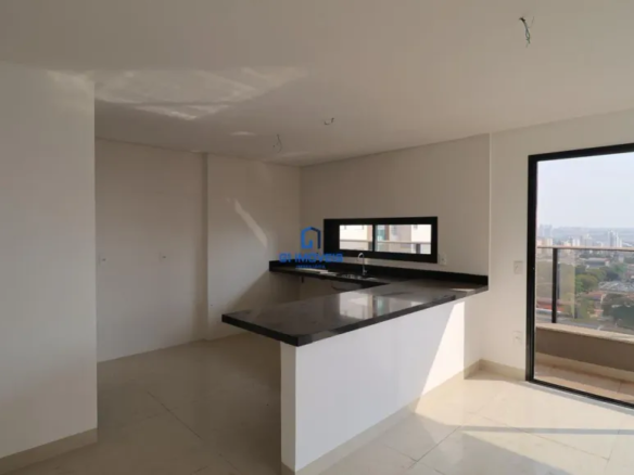 A imagem retrata um interior de um imóvel que parece ser um apartamento ou uma casa moderna. O espaço é luminoso e parece ser uma cozinha integrada com uma área de estar ou jantar, dando uma sensação de amplitude. Cores: As paredes são pintadas em um tom de branco ou bege muito claro, o que contribui para a luminosidade do ambiente. O piso é revestido com cerâmica em um tom claro, possivelmente creme ou bege, complementando a paleta de cores neutras do espaço. A bancada da cozinha é feita de um material escuro, possivelmente granito ou mármore preto, criando um contraste elegante com o restante do ambiente. As esquadrias da janela e da porta que dá acesso à varanda são pretas, proporcionando um toque contemporâneo. Mobiliário: A principal característica do mobiliário é a bancada da cozinha, que se estende em formato de "L". Ela possui uma parte que serve como área de trabalho, com uma pia de aço inoxidável e torneira, e outra parte que se projeta como uma península, funcionando como um balcão para refeições ou um espaço de convivência. A bancada é sustentada por uma base branca, que se harmoniza com as paredes. Ambiente: O ambiente parece estar recém-construído ou reformado, pois está limpo e com um aspecto novo. Há uma janela horizontal acima da área da pia, que permite a entrada de luz natural e oferece uma vista da área externa. Uma porta de vidro deslizante dá acesso a uma varanda, que oferece uma vista panorâmica da cidade, com prédios e vegetação visíveis ao fundo. O teto é liso e branco, com alguns pontos de fiação expostos, sugerindo que a iluminação ainda não foi instalada. Outros Detalhes: Há o logotipo de uma imobiliária ou empresa do setor imobiliário sobreposto na imagem, indicando que o imóvel está sendo promovido para venda ou aluguel. A varanda possui um parapeito de vidro, que permite apreciar a vista sem obstruções. O ambiente parece ser parte de um empreendimento de alto padrão, dada a qualidade dos materiais e o design moderno.