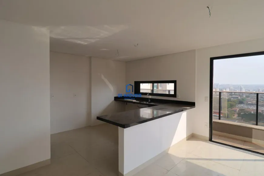 A imagem retrata um interior de um imóvel que parece ser um apartamento ou uma casa moderna. O espaço é luminoso e parece ser uma cozinha integrada com uma área de estar ou jantar, dando uma sensação de amplitude. Cores: As paredes são pintadas em um tom de branco ou bege muito claro, o que contribui para a luminosidade do ambiente. O piso é revestido com cerâmica em um tom claro, possivelmente creme ou bege, complementando a paleta de cores neutras do espaço. A bancada da cozinha é feita de um material escuro, possivelmente granito ou mármore preto, criando um contraste elegante com o restante do ambiente. As esquadrias da janela e da porta que dá acesso à varanda são pretas, proporcionando um toque contemporâneo. Mobiliário: A principal característica do mobiliário é a bancada da cozinha, que se estende em formato de "L". Ela possui uma parte que serve como área de trabalho, com uma pia de aço inoxidável e torneira, e outra parte que se projeta como uma península, funcionando como um balcão para refeições ou um espaço de convivência. A bancada é sustentada por uma base branca, que se harmoniza com as paredes. Ambiente: O ambiente parece estar recém-construído ou reformado, pois está limpo e com um aspecto novo. Há uma janela horizontal acima da área da pia, que permite a entrada de luz natural e oferece uma vista da área externa. Uma porta de vidro deslizante dá acesso a uma varanda, que oferece uma vista panorâmica da cidade, com prédios e vegetação visíveis ao fundo. O teto é liso e branco, com alguns pontos de fiação expostos, sugerindo que a iluminação ainda não foi instalada. Outros Detalhes: Há o logotipo de uma imobiliária ou empresa do setor imobiliário sobreposto na imagem, indicando que o imóvel está sendo promovido para venda ou aluguel. A varanda possui um parapeito de vidro, que permite apreciar a vista sem obstruções. O ambiente parece ser parte de um empreendimento de alto padrão, dada a qualidade dos materiais e o design moderno.