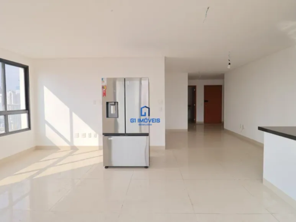 A imagem mostra um ambiente interno amplo e iluminado, possivelmente um apartamento, com as paredes pintadas de branco. A luz natural entra por uma grande janela de moldura preta à esquerda, proporcionando uma vista da cidade. No centro da imagem, destaca-se uma geladeira moderna de aço inoxidável, com um dispenser de água e gelo. Um logotipo da "G1 Imóveis" está sobreposto na geladeira. O chão é revestido com um piso claro e brilhante que reflete a luz, e o teto é branco e sem adornos, com a exceção de algumas instalações elétricas aparentes. À direita, há uma bancada com tampo preto e base branca, sugerindo a área da cozinha. Ao fundo, uma porta de madeira de cor marrom avermelhada indica a entrada para outros cômodos. O ambiente transmite uma sensação de espaço e modernidade, com uma paleta de cores neutras e limpas.