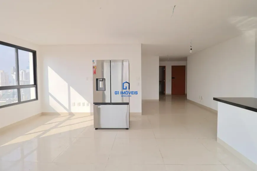 A imagem mostra um ambiente interno amplo e iluminado, possivelmente um apartamento, com as paredes pintadas de branco. A luz natural entra por uma grande janela de moldura preta à esquerda, proporcionando uma vista da cidade. No centro da imagem, destaca-se uma geladeira moderna de aço inoxidável, com um dispenser de água e gelo. Um logotipo da "G1 Imóveis" está sobreposto na geladeira. O chão é revestido com um piso claro e brilhante que reflete a luz, e o teto é branco e sem adornos, com a exceção de algumas instalações elétricas aparentes. À direita, há uma bancada com tampo preto e base branca, sugerindo a área da cozinha. Ao fundo, uma porta de madeira de cor marrom avermelhada indica a entrada para outros cômodos. O ambiente transmite uma sensação de espaço e modernidade, com uma paleta de cores neutras e limpas.