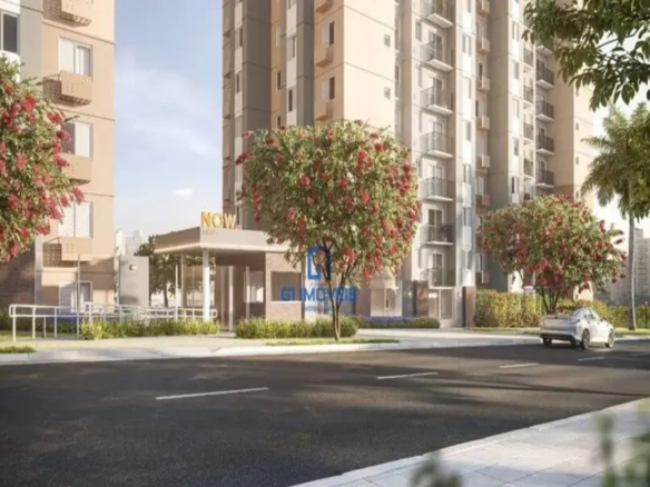 A imagem exibe uma vista da rua de um moderno complexo residencial, possivelmente o now milão. No primeiro plano, uma rua de asfalto escura com faixas brancas tracejadas ocupa a parte inferior da cena, ladeada por uma calçada de blocos claros e um trecho de grama. Elevando-se ao fundo, estão vários edifícios de apartamentos de múltiplos andares. As fachadas combinam tons de bege claro, cinza e detalhes em branco, com numerosas janelas e varandas. Alguns apartamentos possuem unidades de ar condicionado externas visíveis. No centro-esquerda, destaca-se a entrada do complexo, uma estrutura coberta com design contemporâneo, paredes que parecem ser de vidro ou painéis claros, e um letreiro proeminente em letras douradas ou amarelas onde se lê "NOW" e, abaixo, em letras menores, provavelmente o complemento do nome do empreendimento. Uma rampa de acesso leva à entrada. O paisagismo é bem cuidado, com gramados verdes e arbustos baixos. Diversas árvores floridas, com profusas flores em tons de vermelho-rosado, adornam a calçada e as áreas verdes, adicionando cor vibrante à cena. À direita, um carro sedan prateado trafega pela rua, afastando-se do observador. Ao fundo, mais vegetação, incluindo o que parecem ser palmeiras, e a silhueta de outros edifícios urbanos podem ser vistos sob um céu claro e luminoso, sugerindo um dia ensolarado.
