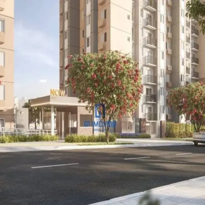 A imagem exibe uma vista da rua de um moderno complexo residencial, possivelmente o now milão. No primeiro plano, uma rua de asfalto escura com faixas brancas tracejadas ocupa a parte inferior da cena, ladeada por uma calçada de blocos claros e um trecho de grama. Elevando-se ao fundo, estão vários edifícios de apartamentos de múltiplos andares. As fachadas combinam tons de bege claro, cinza e detalhes em branco, com numerosas janelas e varandas. Alguns apartamentos possuem unidades de ar condicionado externas visíveis. No centro-esquerda, destaca-se a entrada do complexo, uma estrutura coberta com design contemporâneo, paredes que parecem ser de vidro ou painéis claros, e um letreiro proeminente em letras douradas ou amarelas onde se lê "NOW" e, abaixo, em letras menores, provavelmente o complemento do nome do empreendimento. Uma rampa de acesso leva à entrada. O paisagismo é bem cuidado, com gramados verdes e arbustos baixos. Diversas árvores floridas, com profusas flores em tons de vermelho-rosado, adornam a calçada e as áreas verdes, adicionando cor vibrante à cena. À direita, um carro sedan prateado trafega pela rua, afastando-se do observador. Ao fundo, mais vegetação, incluindo o que parecem ser palmeiras, e a silhueta de outros edifícios urbanos podem ser vistos sob um céu claro e luminoso, sugerindo um dia ensolarado.