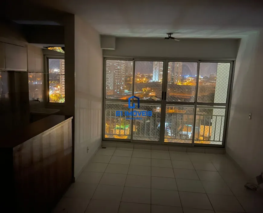 A imagem retrata o interior de um apartamento durante a noite, focando em um espaço que integra cozinha e sala. A iluminação é predominantemente artificial, com o brilho da cidade visível através de uma grande janela dominando o ambiente. Cozinha (Lado Esquerdo): Armários: Os armários são construídos em madeira escura, possivelmente mogno ou um verniz similar. O acabamento parece ser liso, com um certo brilho que reflete a luz. As portas dos armários são de design simples e reto. Pia: A pia é de cerâmica branca ou um material similar de cor clara. A torneira é de metal, provavelmente cromada, e reflete a luz. Há um respingo de azulejos na parede atrás da pia, que parece ser de cor clara e pode ter um padrão discreto. Bancada: Há uma bancada de madeira mais clara, possivelmente pinho ou uma madeira similar, que serve de divisória entre a cozinha e a sala. A madeira da bancada parece ter uma coloração mais clara que os armários. Sala de Estar/Quarto (Lado Direito): Paredes: As paredes são pintadas em um tom claro de branco ou bege. A superfície parece lisa e sem adornos, com exceção de uma tomada ou interruptor visível em uma das paredes. Janela: A janela é o ponto focal do ambiente. É uma grande janela de correr com várias divisórias de metal, provavelmente alumínio pintado de branco ou cinza claro. Há uma grade de proteção de metal do lado de fora da janela, adicionando uma camada de segurança. Uma tela mosquiteira também está presente, indicando a preocupação com insetos. Vista: A vista da janela é um panorama urbano noturno. Edifícios altos com muitas janelas iluminadas se estendem até onde a vista alcança. Há luzes de carros nas ruas e avenidas, criando rastros luminosos. A iluminação da cidade é intensa, contrastando com o céu noturno escuro. Piso: O piso é revestido com azulejos quadrados de cor clara, provavelmente cerâmica ou porcelanato. As juntas entre os azulejos são visíveis, criando um padrão quadriculado. O piso parece limpo e bem conservado. Iluminação: A iluminação interna é fornecida por uma luminária de teto discreta, que parece ser um modelo simples e funcional. A luz é suave e difusa, iluminando o ambiente de maneira uniforme. Detalhes adicionais: É possível notar um objeto no chão, próximo a parede da direita, que pode ser um saco plástico, ou um objeto não identificado. Atmosfera Geral: A atmosfera geral do ambiente é de calma e tranquilidade, com a iluminação suave e a vista da cidade criando um cenário relaxante. A simplicidade do design e a funcionalidade dos móveis sugerem um estilo de vida prático e descomplicado.