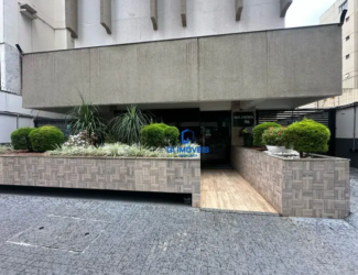 Visão Geral: Estilo Arquitetônico: O edifício apresenta um estilo modernista com ênfase em linhas retas e superfícies planas. Há uma forte influência do concreto aparente, tanto na marquise quanto nos elementos decorativos da fachada. Iluminação: A cena parece ser iluminada por luz natural difusa, sugerindo um dia nublado ou uma área com sombra parcial. Composição: A imagem é bem composta, com a entrada do edifício no centro e os elementos paisagísticos em ambos os lados, criando um senso de equilíbrio e convidando o espectador a entrar. Detalhes da Entrada: Piso: O piso externo é feito de ladrilhos cinzas com um padrão que lembra paralelepípedos. As juntas entre os ladrilhos são estreitas e bem definidas. Rampa: A rampa de acesso é revestida com madeira clara, possivelmente tratada para resistir às intempéries. As tábuas são dispostas longitudinalmente e parecem ter um acabamento liso. Canteiros: Os canteiros são elevados e revestidos com ladrilhos retangulares em tons de bege e cinza. Os ladrilhos são dispostos em um padrão complexo que simula um mosaico ou um cesto de vime. A borda superior dos canteiros é bem definida e reta. Vegetação: Arbustos: Os arbustos são densos e arredondados, com folhagem verde escura brilhante. Eles parecem ser podados regularmente para manter sua forma. Gramíneas Ornamentais: As gramíneas têm folhas longas e finas que se movem suavemente com o vento. Elas adicionam textura e movimento aos canteiros. Flores: As flores brancas são pequenas e delicadas, com pétalas finas e centros amarelos. Elas contrastam com a folhagem escura e adicionam um toque de cor aos canteiros. Vasos: Os vasos brancos são redondos e simples, com bordas largas e curvas. Eles são preenchidos com plantas de folhagem verde escura e cobertos com pedras brancas para reter a umidade e evitar o crescimento de ervas daninhas. Porta de Entrada: A porta de entrada é feita de vidro escuro, possivelmente temperado para maior segurança. Ela reflete a luz e o ambiente ao redor, criando um efeito interessante. Uma placa preta acima da porta exibe o nome do edifício ("SOLIMÕES") e o número ("96") em letras brancas. Detalhes Adicionais: Há uma placa ou adesivo com o logotipo de uma imobiliária (GL IMOVEIS) sobreposta à imagem, indicando que o edifício está sendo comercializado ou administrado por essa empresa. Detalhes da Fachada: Marquise: A marquise é feita de concreto aparente com uma textura áspera e irregular. Ela se projeta para fora da fachada, criando uma área sombreada sobre a entrada. Revestimento: O revestimento da fachada é feito de um material de cor clara, possivelmente concreto ou alvenaria pintada. Ele tem uma textura lisa e uniforme. Elementos Decorativos: Os elementos decorativos em concreto aparente adicionam interesse visual à fachada. Eles têm formas geométricas simples e se projetam para fora da parede. Ambiente: Urbano: A imagem sugere um ambiente urbano, com edifícios altos e ruas movimentadas. Cuidado: O edifício parece ser bem cuidado e mantido, com a vegetação podada e a limpeza em dia. Acessibilidade: A rampa de acesso indica que o edifício é acessível a pessoas com mobilidade reduzida.