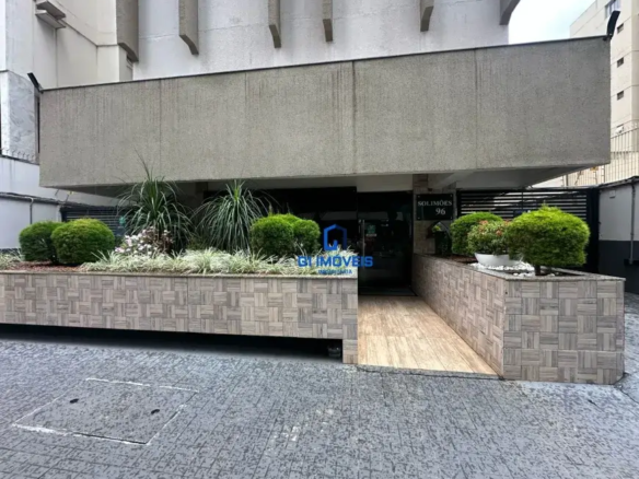 Visão Geral: Estilo Arquitetônico: O edifício apresenta um estilo modernista com ênfase em linhas retas e superfícies planas. Há uma forte influência do concreto aparente, tanto na marquise quanto nos elementos decorativos da fachada. Iluminação: A cena parece ser iluminada por luz natural difusa, sugerindo um dia nublado ou uma área com sombra parcial. Composição: A imagem é bem composta, com a entrada do edifício no centro e os elementos paisagísticos em ambos os lados, criando um senso de equilíbrio e convidando o espectador a entrar. Detalhes da Entrada: Piso: O piso externo é feito de ladrilhos cinzas com um padrão que lembra paralelepípedos. As juntas entre os ladrilhos são estreitas e bem definidas. Rampa: A rampa de acesso é revestida com madeira clara, possivelmente tratada para resistir às intempéries. As tábuas são dispostas longitudinalmente e parecem ter um acabamento liso. Canteiros: Os canteiros são elevados e revestidos com ladrilhos retangulares em tons de bege e cinza. Os ladrilhos são dispostos em um padrão complexo que simula um mosaico ou um cesto de vime. A borda superior dos canteiros é bem definida e reta. Vegetação: Arbustos: Os arbustos são densos e arredondados, com folhagem verde escura brilhante. Eles parecem ser podados regularmente para manter sua forma. Gramíneas Ornamentais: As gramíneas têm folhas longas e finas que se movem suavemente com o vento. Elas adicionam textura e movimento aos canteiros. Flores: As flores brancas são pequenas e delicadas, com pétalas finas e centros amarelos. Elas contrastam com a folhagem escura e adicionam um toque de cor aos canteiros. Vasos: Os vasos brancos são redondos e simples, com bordas largas e curvas. Eles são preenchidos com plantas de folhagem verde escura e cobertos com pedras brancas para reter a umidade e evitar o crescimento de ervas daninhas. Porta de Entrada: A porta de entrada é feita de vidro escuro, possivelmente temperado para maior segurança. Ela reflete a luz e o ambiente ao redor, criando um efeito interessante. Uma placa preta acima da porta exibe o nome do edifício ("SOLIMÕES") e o número ("96") em letras brancas. Detalhes Adicionais: Há uma placa ou adesivo com o logotipo de uma imobiliária (GL IMOVEIS) sobreposta à imagem, indicando que o edifício está sendo comercializado ou administrado por essa empresa. Detalhes da Fachada: Marquise: A marquise é feita de concreto aparente com uma textura áspera e irregular. Ela se projeta para fora da fachada, criando uma área sombreada sobre a entrada. Revestimento: O revestimento da fachada é feito de um material de cor clara, possivelmente concreto ou alvenaria pintada. Ele tem uma textura lisa e uniforme. Elementos Decorativos: Os elementos decorativos em concreto aparente adicionam interesse visual à fachada. Eles têm formas geométricas simples e se projetam para fora da parede. Ambiente: Urbano: A imagem sugere um ambiente urbano, com edifícios altos e ruas movimentadas. Cuidado: O edifício parece ser bem cuidado e mantido, com a vegetação podada e a limpeza em dia. Acessibilidade: A rampa de acesso indica que o edifício é acessível a pessoas com mobilidade reduzida.