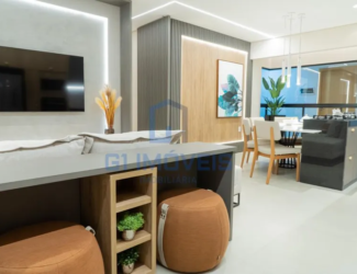 A imagem mostra um espaço interno moderno e bem iluminado, provavelmente a sala de estar e jantar de um apartamento ou casa. As cores predominantes são tons neutros como cinza, branco e madeira clara, com toques de cor em elementos decorativos. Na parede da esquerda, há uma televisão de tela plana pendurada em uma parede texturizada cinza, com uma faixa de luz LED vertical iluminando a lateral. Em frente à TV, parte de um sofá cinza claro pode ser vista, sugerindo uma área de estar confortável. Na parte inferior da imagem, vemos uma bancada cinza escura com um design funcional. Ela possui um nicho de madeira clara com prateleiras, onde pequenos vasos com plantas verdes estão dispostos. Ao lado, há dois pufes marrons com detalhes em alças claras, adicionando assentos extras e um toque casual ao ambiente. Ao fundo, a parede é revestida com painéis verticais cinza escuro, contrastando com uma faixa de madeira clara no centro, onde um quadro com motivos botânicos em tons de azul e verde está pendurado. A área de jantar é visível ao fundo, com uma mesa redonda branca e cadeiras de madeira clara com estofamento branco. Luminárias pendentes finas e delicadas iluminam a mesa. Uma planta alta em um vaso adiciona um toque de natureza ao espaço. Uma divisória de vidro separa a cozinha da área de jantar, com um balcão embutido que parece ter um fogão cooktop. O design geral é limpo e minimalista, com linhas retas e uma paleta de cores suaves, criando um ambiente acolhedor e sofisticado.