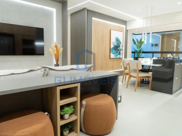 A imagem mostra um espaço interno moderno e bem iluminado, provavelmente a sala de estar e jantar de um apartamento ou casa. As cores predominantes são tons neutros como cinza, branco e madeira clara, com toques de cor em elementos decorativos. Na parede da esquerda, há uma televisão de tela plana pendurada em uma parede texturizada cinza, com uma faixa de luz LED vertical iluminando a lateral. Em frente à TV, parte de um sofá cinza claro pode ser vista, sugerindo uma área de estar confortável. Na parte inferior da imagem, vemos uma bancada cinza escura com um design funcional. Ela possui um nicho de madeira clara com prateleiras, onde pequenos vasos com plantas verdes estão dispostos. Ao lado, há dois pufes marrons com detalhes em alças claras, adicionando assentos extras e um toque casual ao ambiente. Ao fundo, a parede é revestida com painéis verticais cinza escuro, contrastando com uma faixa de madeira clara no centro, onde um quadro com motivos botânicos em tons de azul e verde está pendurado. A área de jantar é visível ao fundo, com uma mesa redonda branca e cadeiras de madeira clara com estofamento branco. Luminárias pendentes finas e delicadas iluminam a mesa. Uma planta alta em um vaso adiciona um toque de natureza ao espaço. Uma divisória de vidro separa a cozinha da área de jantar, com um balcão embutido que parece ter um fogão cooktop. O design geral é limpo e minimalista, com linhas retas e uma paleta de cores suaves, criando um ambiente acolhedor e sofisticado.