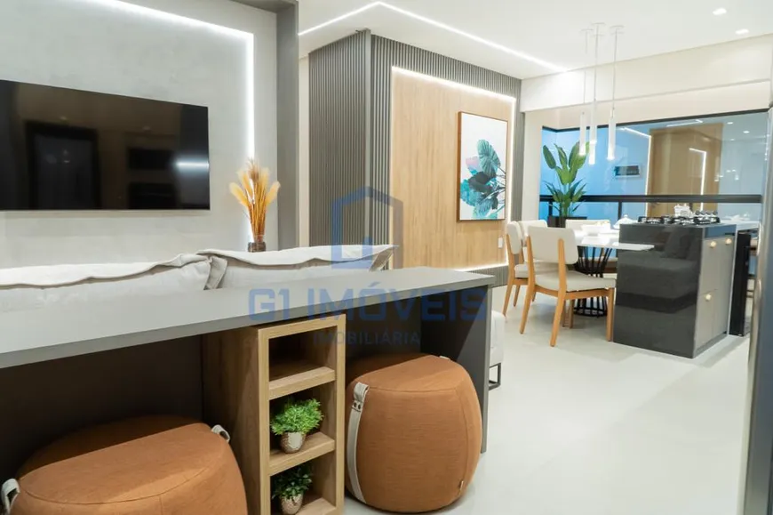 A imagem mostra um espaço interno moderno e bem iluminado, provavelmente a sala de estar e jantar de um apartamento ou casa. As cores predominantes são tons neutros como cinza, branco e madeira clara, com toques de cor em elementos decorativos. Na parede da esquerda, há uma televisão de tela plana pendurada em uma parede texturizada cinza, com uma faixa de luz LED vertical iluminando a lateral. Em frente à TV, parte de um sofá cinza claro pode ser vista, sugerindo uma área de estar confortável. Na parte inferior da imagem, vemos uma bancada cinza escura com um design funcional. Ela possui um nicho de madeira clara com prateleiras, onde pequenos vasos com plantas verdes estão dispostos. Ao lado, há dois pufes marrons com detalhes em alças claras, adicionando assentos extras e um toque casual ao ambiente. Ao fundo, a parede é revestida com painéis verticais cinza escuro, contrastando com uma faixa de madeira clara no centro, onde um quadro com motivos botânicos em tons de azul e verde está pendurado. A área de jantar é visível ao fundo, com uma mesa redonda branca e cadeiras de madeira clara com estofamento branco. Luminárias pendentes finas e delicadas iluminam a mesa. Uma planta alta em um vaso adiciona um toque de natureza ao espaço. Uma divisória de vidro separa a cozinha da área de jantar, com um balcão embutido que parece ter um fogão cooktop. O design geral é limpo e minimalista, com linhas retas e uma paleta de cores suaves, criando um ambiente acolhedor e sofisticado.