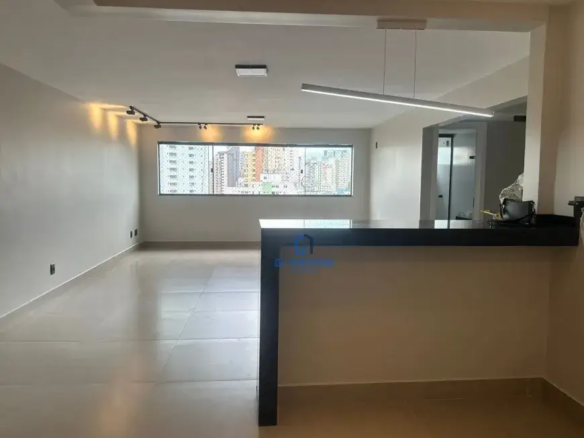 Estrutura e Acabamentos: Teto: O teto apresenta um rebaixamento em gesso, criando um efeito de moldura que delimita o espaço e valoriza a iluminação. A luminária retangular suspensa por cabos finos, provavelmente de aço, emite uma luz branca e fria que se espalha uniformemente pelo ambiente. Paredes: As paredes, em um tom de bege muito claro, quase um off-white, possuem um acabamento liso e uniforme. Próximo ao piso, observa-se um rodapé discreto, também em tom claro, que acompanha toda a extensão das paredes. Há algumas tomadas e interruptores instalados, indicando a infraestrutura elétrica do apartamento. Piso: O piso é revestido por cerâmicas de grande formato, com juntas mínimas, o que contribui para a sensação de amplitude e continuidade visual. O acabamento brilhante reflete a luz e realça a limpeza do ambiente. A cor do piso é um bege suave, um pouco mais escuro que as paredes, criando um contraste sutil e elegante. Janela: A janela panorâmica é composta por várias folhas de vidro, provavelmente com esquadrias de alumínio na cor preta ou grafite. A vista da paisagem urbana revela edifícios de diferentes alturas e cores, com uma predominância de tons claros e alguns detalhes em cores vibrantes. As janelas são translúcidas, com venezianas horizontais. Iluminação: Luz natural: A grande janela permite a entrada abundante de luz natural, que ilumina o ambiente e proporciona uma sensação de bem-estar. A luz natural também destaca as cores e texturas dos materiais utilizados no acabamento do apartamento. Iluminação artificial: O trilho de iluminação com spots direcionáveis oferece uma iluminação mais flexível e personalizável. Os spots podem ser ajustados para destacar objetos de decoração, quadros ou áreas específicas do ambiente. A luminária retangular suspensa complementa a iluminação geral do espaço, proporcionando uma luz suave e difusa. Bancada Divisória: Estrutura: A bancada divisória é composta por uma base em alvenaria revestida com o mesmo tom de bege das paredes, e um tampo em granito preto, com bordas retas e acabamento polido. A bancada possui uma altura padrão, que permite o uso de banquetas para refeições rápidas ou para socializar com amigos e familiares. Detalhes: Sobre a bancada, observa-se uma bolsa preta de couro, com alças e detalhes em metal, que sugere que o espaço está sendo preparado para ser ocupado. Também é possível ver outros objetos, como um pote com alguns talheres em cima, provavelmente decorativos, e uma garrafa de água ou suco. Outros Detalhes: Porta: Ao lado da bancada, é possível ver uma porta branca, provavelmente que dá acesso a um lavabo ou banheiro social. A porta possui um design clean e minimalista, com puxador discreto e acabamento em pintura lisa. Infraestrutura: A presença de tomadas e interruptores nas paredes indica que o apartamento possui uma infraestrutura elétrica completa, com pontos de energia e iluminação estrategicamente distribuídos. Sensação Geral: A imagem transmite uma sensação de modernidade, sofisticação e bom gosto. A combinação de cores claras, a iluminação abundante e o acabamento impecável criam um ambiente agradável e convidativo, perfeito para quem busca um lar confortável e elegante. A falta de móveis permite que o futuro morador personalize o espaço de acordo com suas preferências, tornando-o único e especial.