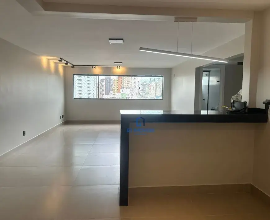 Estrutura e Acabamentos: Teto: O teto apresenta um rebaixamento em gesso, criando um efeito de moldura que delimita o espaço e valoriza a iluminação. A luminária retangular suspensa por cabos finos, provavelmente de aço, emite uma luz branca e fria que se espalha uniformemente pelo ambiente. Paredes: As paredes, em um tom de bege muito claro, quase um off-white, possuem um acabamento liso e uniforme. Próximo ao piso, observa-se um rodapé discreto, também em tom claro, que acompanha toda a extensão das paredes. Há algumas tomadas e interruptores instalados, indicando a infraestrutura elétrica do apartamento. Piso: O piso é revestido por cerâmicas de grande formato, com juntas mínimas, o que contribui para a sensação de amplitude e continuidade visual. O acabamento brilhante reflete a luz e realça a limpeza do ambiente. A cor do piso é um bege suave, um pouco mais escuro que as paredes, criando um contraste sutil e elegante. Janela: A janela panorâmica é composta por várias folhas de vidro, provavelmente com esquadrias de alumínio na cor preta ou grafite. A vista da paisagem urbana revela edifícios de diferentes alturas e cores, com uma predominância de tons claros e alguns detalhes em cores vibrantes. As janelas são translúcidas, com venezianas horizontais. Iluminação: Luz natural: A grande janela permite a entrada abundante de luz natural, que ilumina o ambiente e proporciona uma sensação de bem-estar. A luz natural também destaca as cores e texturas dos materiais utilizados no acabamento do apartamento. Iluminação artificial: O trilho de iluminação com spots direcionáveis oferece uma iluminação mais flexível e personalizável. Os spots podem ser ajustados para destacar objetos de decoração, quadros ou áreas específicas do ambiente. A luminária retangular suspensa complementa a iluminação geral do espaço, proporcionando uma luz suave e difusa. Bancada Divisória: Estrutura: A bancada divisória é composta por uma base em alvenaria revestida com o mesmo tom de bege das paredes, e um tampo em granito preto, com bordas retas e acabamento polido. A bancada possui uma altura padrão, que permite o uso de banquetas para refeições rápidas ou para socializar com amigos e familiares. Detalhes: Sobre a bancada, observa-se uma bolsa preta de couro, com alças e detalhes em metal, que sugere que o espaço está sendo preparado para ser ocupado. Também é possível ver outros objetos, como um pote com alguns talheres em cima, provavelmente decorativos, e uma garrafa de água ou suco. Outros Detalhes: Porta: Ao lado da bancada, é possível ver uma porta branca, provavelmente que dá acesso a um lavabo ou banheiro social. A porta possui um design clean e minimalista, com puxador discreto e acabamento em pintura lisa. Infraestrutura: A presença de tomadas e interruptores nas paredes indica que o apartamento possui uma infraestrutura elétrica completa, com pontos de energia e iluminação estrategicamente distribuídos. Sensação Geral: A imagem transmite uma sensação de modernidade, sofisticação e bom gosto. A combinação de cores claras, a iluminação abundante e o acabamento impecável criam um ambiente agradável e convidativo, perfeito para quem busca um lar confortável e elegante. A falta de móveis permite que o futuro morador personalize o espaço de acordo com suas preferências, tornando-o único e especial.