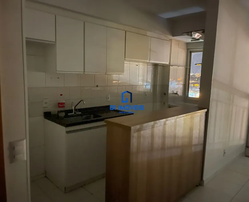 Visão Geral: A imagem captura uma cozinha compacta e funcional, projetada em um estilo moderno e prático. A iluminação é suave e difusa, criando uma atmosfera convidativa e aconchegante. A composição da imagem foca principalmente na área da pia e do balcão, oferecendo uma visão detalhada dos materiais e acessórios utilizados. Detalhes da Parede e Azulejos: As paredes são revestidas com azulejos quadrados de cor creme claro, quase branco. As juntas entre os azulejos são finas e claras, proporcionando uma aparência limpa e uniforme. Há algumas pequenas imperfeições e marcas sutis nos azulejos, sugerindo que a cozinha é utilizada e bem cuidada. Armários Superiores: Os armários superiores são brancos e possuem um acabamento liso e brilhante. As portas são retangulares e sem detalhes ornamentais, com puxadores discretos em metal prateado. A parte superior dos armários acompanha a linha do teto, otimizando o espaço de armazenamento vertical. Bancada e Pia: A bancada é feita de granito preto com pequenos pontos de brilho, conferindo um toque de sofisticação. A pia é de aço inoxidável e possui um design retangular, com cantos arredondados. A torneira é moderna, com um corpo cromado e um design ergonômico. Ao lado da pia, uma garrafa de Coca-Cola vermelha adiciona uma pitada de cor e um toque casual ao ambiente. Balcão de Madeira: O balcão de madeira serve como uma barreira visual e funcional entre a cozinha e outro espaço adjacente. A madeira possui um tom claro e natural, com um acabamento que realça os veios e a textura da madeira. A estrutura do balcão é simples, com painéis verticais que se estendem do chão até a altura da bancada. Iluminação e Vista: A iluminação na cozinha é suave e indireta, criando uma atmosfera agradável. A luz natural entra pela janela, que oferece uma vista noturna da cidade. As luzes da cidade criam um cenário cintilante e colorido, que contrasta com a sobriedade da cozinha. Detalhes Adicionais: Na parede, perto da pia, há uma tomada elétrica com um espelho branco. O piso da cozinha é revestido com cerâmica clara, que complementa a paleta de cores do ambiente. Há uma pequena abertura entre o balcão e a parede, revelando parte do espaço adjacente. A imagem é bem iluminada e possui uma boa profundidade de campo, permitindo que os detalhes sejam vistos com clareza. Em resumo, a cozinha é um espaço funcional e bem projetado, com uma estética moderna e convidativa. A combinação de cores claras, materiais de qualidade e iluminação suave cria um ambiente agradável e acolhedor.