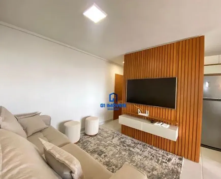 A imagem apresenta um espaço de convivência moderno e convidativo, caracterizado por uma paleta de cores neutras e materiais contrastantes. A iluminação é uniforme e suave, destacando os detalhes dos móveis e da decoração. Paredes e Iluminação: As paredes são pintadas em um branco suave, possivelmente um tom off-white, que reflete a luz natural e artificial. Há um rodapé branco fino que percorre toda a extensão da parede, criando uma separação limpa entre a parede e o piso. No teto, uma luminária quadrada embutida emite uma luz difusa e uniforme, iluminando toda a área. Mobiliário: Sofá: O sofá é do tipo secional, com um tom bege claro que se harmoniza com o restante da decoração. Ele possui almofadas macias e convidativas, com diferentes tamanhos e texturas. Pufes: Dois pufes cilíndricos brancos servem como assentos adicionais ou apoio para os pés. Eles têm um acabamento liso e clean. Rack: O rack flutuante abaixo da TV é branco e possui um design minimalista. Ele oferece espaço para acomodar aparelhos eletrônicos, como um roteador Wi-Fi (visível pelos cabos conectados) e outros dispositivos. Detalhes de Decoração e Acabamento: Parede de Madeira: A parede de madeira vertical é um elemento marcante na sala. Ela é composta por ripas de madeira estreitas e verticais, com um tom marrom-médio que adiciona calor e textura ao espaço. A TV de tela plana está fixada nesta parede, criando um ponto focal. Tapete: O tapete é um dos elementos mais texturizados da sala. Ele possui um padrão abstrato em tons de cinza e branco, com um visual desgastado ou sutilmente manchado. Suas fibras parecem macias e aconchegantes. Cozinha: No fundo, a cozinha apresenta uma geladeira de aço inoxidável com um acabamento escovado. Os armários são brancos e têm um design moderno e clean. Uma pequena parte da bancada também é visível. Porta: Ao lado da parede de madeira, uma porta de madeira maciça em tom marrom-médio oferece acesso a outra área do apartamento. A porta tem um design simples e funcional. Atmosfera Geral: A sala de estar transmite uma sensação de conforto, organização e modernidade. A combinação de cores neutras, materiais naturais (como a madeira) e elementos de design minimalistas cria um ambiente agradável e convidativo. A iluminação suave e a disposição dos móveis favorecem o relaxamento e a convivência.