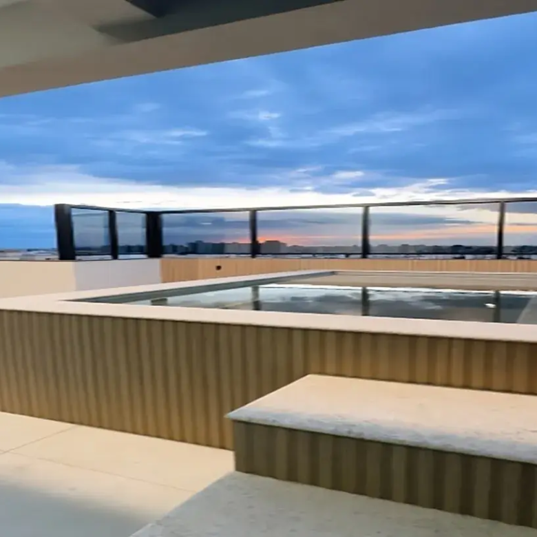 Aqui está uma descrição da imagem: A imagem mostra uma piscina em uma cobertura com vista para um horizonte urbano ao pôr do sol. As cores são dominadas por tons de azul e laranja do céu e pelos tons neutros da piscina e do deque. A piscina é retangular e cercada por uma parede de madeira clara. Uma borda de concreto branco percorre o topo da parede da piscina e um banco de concreto fica na frente da piscina. O piso é de cerâmica clara e contribui para a sensação de limpeza e modernidade. Ao fundo, um horizonte urbano com prédios de vários tamanhos pode ser visto. A vista é parcialmente obstruída por uma barreira de vidro preta que circunda o deck da cobertura. O céu está nublado e o sol está se pondo, criando um brilho laranja no horizonte. A cena geral é de luxo e sofisticação. A piscina e o deque da cobertura oferecem um espaço relaxante e elegante com uma vista deslumbrante da cidade. A paleta de cores é relaxante e convidativa, e os materiais utilizados são de alta qualidade. Palavras-chave: Piscina, cobertura, cidade, horizonte, pôr do sol, luxo, sofisticação, relaxante, elegante, materiais de alta qualidade. cobertura a venda em uberlândia Palavras chave: Cobertura duplex com 4 quartos à venda 185m² no Santa Mônica Cobertura duplex com 4 quartos à venda 180m² no Santa Mônica