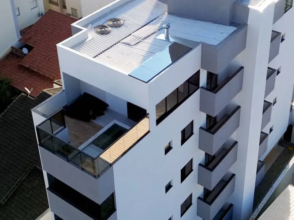 Foco na Fachada e Cobertura do Edifício Meraki, Santa Mônica, Uberlândia: 1. Visão Geral da Imagem: Perspectiva: A imagem nos presenteia com uma visão aérea e angular do Edifício Meraki, permitindo uma análise minuciosa da sua fachada e cobertura. O ângulo escolhido enfatiza a verticalidade do edifício e a relação entre seus elementos arquitetônicos. Enquadramento: O enquadramento da imagem concentra-se na parte superior do edifício, proporcionando uma visão clara e detalhada da sua estrutura, materiais e acabamentos. 2. Fachada: Uma Expressão de Modernidade e Elegância: Design: A fachada do Edifício Meraki expressa um design moderno e minimalista, com linhas retas e volumes geométricos bem definidos. A ausência de elementos decorativos excessivos enfatiza a pureza das formas e a elegância do conjunto. Cores: A paleta de cores da fachada é composta por tons neutros e suaves, como branco e cinza. O branco confere luminosidade e amplitude ao edifício, enquanto o cinza adiciona um toque de sofisticação e sobriedade. As janelas escuras criam um contraste marcante com os painéis claros, acentuando a verticalidade da construção. Sacadas: As sacadas do Edifício Meraki são integradas à fachada, com um design discreto e elegante. Os guarda-corpos de vidro proporcionam segurança e privacidade aos moradores, sem comprometer a vista da cidade. As sacadas criam uma sensação de continuidade entre o interior e o exterior dos apartamentos, ampliando os espaços e permitindo o contato com a natureza. Janelas: As janelas retangulares da fachada são amplas e generosas, permitindo a entrada abundante de luz natural nos apartamentos. As molduras escuras das janelas criam um contraste elegante com os painéis claros da fachada, acentuando a verticalidade do edifício. Painéis: A fachada do Edifício Meraki é composta por painéis pré-fabricados, que garantem a qualidade, a durabilidade e a rapidez da construção. Os painéis apresentam um acabamento liso e uniforme, conferindo um aspecto moderno e sofisticado ao edifício. 3. Cobertura: Um Espaço de Lazer e Bem-Estar: Design: A cobertura do Edifício Meraki apresenta um design inovador e funcional, com diferentes níveis e espaços de convivência. A área de lazer na cobertura é um oásis de bem-estar e entretenimento, com piscina, solarium, churrasqueira e outros ambientes projetados para o conforto e a diversão dos moradores. Materiais: A cobertura do Edifício Meraki é revestida com materiais de alta qualidade e resistência, como telhas metálicas, mantas impermeabilizantes e porcelanatos antiderrapantes. Os materiais escolhidos garantem a durabilidade, a segurança e a beleza do espaço. Piscina: A piscina na cobertura é um dos destaques do Edifício Meraki, oferecendo um espaço para relaxamento, prática de esportes e convívio social. A água cristalina da piscina reflete o céu e a paisagem urbana, criando uma atmosfera mágica e inspiradora. Telhado: A cobertura telhado do edifício é telhado com telhas metálicas, com um bom sistema de escoamento de aguas pluvias. Equipamentos: A cobertura do Edifício Meraki abriga diversos equipamentos técnicos, como aparelhos de ar condicionado, caixas d'água e chaminés. Os equipamentos são instalados de forma organizada e discreta, sem comprometer a estética do espaço. Vista: A cobertura do Edifício Meraki oferece uma vista privilegiada da cidade de Uberlândia. Os moradores podem desfrutar de paisagens panorâmicas, pores do sol inesquecíveis e momentos de lazer em um ambiente exclusivo e sofisticado. 4. Relação com o Entorno: Integração: A arquitetura do Edifício Meraki busca integrar-se harmoniosamente com o entorno urbano de Santa Mônica. O edifício respeita a escala do bairro, com uma altura que não compromete a vista dos vizinhos e um design que não destoa das construções existentes. Valorização: A construção do Edifício Meraki contribui para a valorização do bairro Santa Mônica, atraindo novos moradores e investidores e impulsionando o desenvolvimento da região. Palavra chave Apartamento à Venda com 3 Quartos 93m² no Setor Santa Mônica