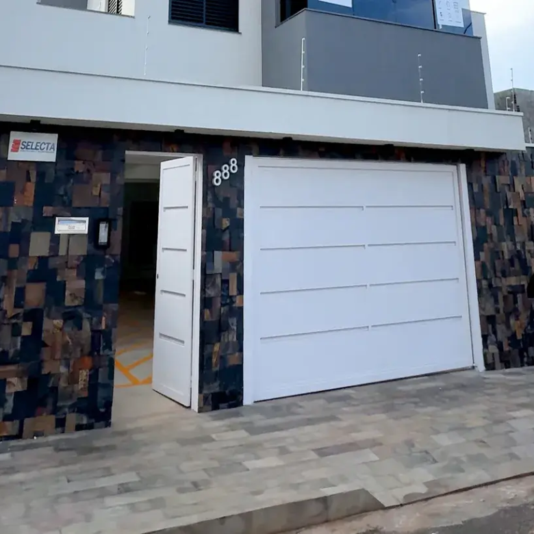 Fachada de imóvel com muro revestido em pedra escura. Destaque para o portão e a porta de entrada brancos, a inscrição "MERAKI" no muro e cerca elétrica de segurança.