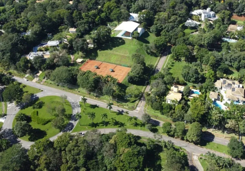 Esta é uma vista aérea do Aldeia do Vale em Goiânia, tirada de um ponto de vista elevado, que captura uma paisagem exuberante e bem desenvolvida característica deste renomado condomínio residencial de alto padrão. No centro da imagem, ligeiramente deslocado para a esquerda, destacam-se duas quadras de tênis de saibro, de cor laranja-avermelhada, perfeitamente demarcadas com linhas brancas e redes pretas. Elas estão dispostas lado a lado e cercadas por uma área gramada e algumas árvores. Pequenas estruturas brancas, possivelmente bancos ou abrigos para jogadores, são visíveis ao lado das quadras. Acima e à direita das quadras de tênis, em um terreno gramado e levemente inclinado, ergue-se uma grande construção com um proeminente telhado branco de múltiplas águas, com um design moderno e angular. Essa estrutura pode ser um ginásio, uma quadra coberta ou um salão de eventos do condomínio. Uma rede de estradas asfaltadas, de cor cinza-escura, serpenteia pela paisagem, conectando as diferentes áreas. No canto inferior esquerdo, há uma rotatória bem definida, com uma ilha central gramada que apresenta um desenho paisagístico distintivo, possivelmente um arbusto podado em formato curvo ou de vírgula. Ao longo de uma das vias que se afasta da rotatória, há uma fileira de palmeiras altas e esguias. Alguns carros, de cor clara, podem ser vistos estacionados ou trafegando pelas vias. A vegetação é um elemento dominante na imagem. Uma vasta extensão de árvores de diferentes espécies e tamanhos preenche a maior parte do quadro, formando uma densa cobertura verde. As árvores circundam as construções, ladeiam as estradas e preenchem os espaços abertos, conferindo à área uma sensação de privacidade e integração com a natureza. Os gramados são bem cuidados e de um verde vibrante. Dispersas entre a vegetação, avistam-se diversas residências. São casas grandes, com arquitetura moderna, predominantemente brancas ou de cores claras. Muitas delas possuem telhados planos ou com pouca inclinação, e algumas apresentam telhados de telha cerâmica marrom. No canto superior direito e no canto inferior direito, conjuntos de casas são mais visíveis, algumas com piscinas de água azul clara e, em certos casos, painéis solares instalados nos telhados. A iluminação sugere um dia ensolarado, com sombras projetadas pelas árvores e construções, indicando breakouts direção da luz solar. A qualidade da imagem é nítida, permitindo a observação de muitos detalhes. A impressão geral é de um ambiente organizado, próspero, tranquilo e com forte presença de áreas verdes bem planejadas, típico do Aldeia do Vale.