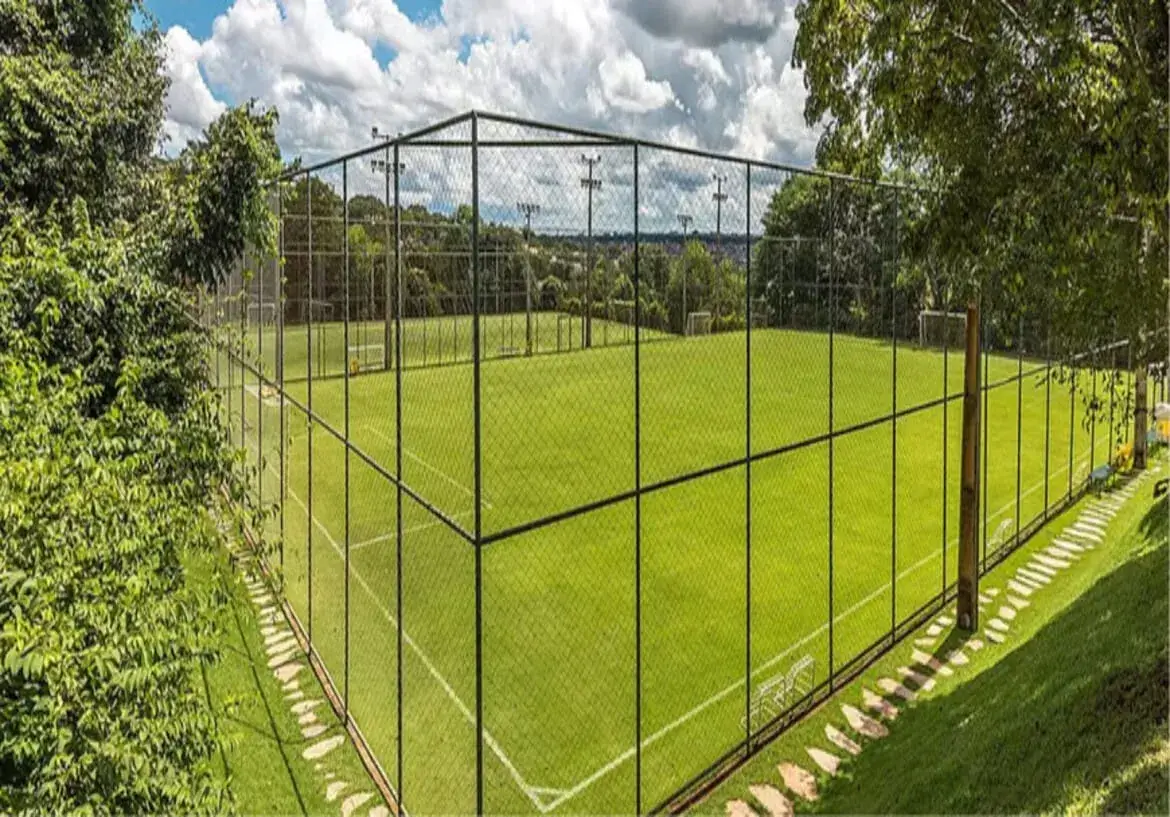 Campo de futebol society no condomínio aldeia do vale com grama sintética, cercado por alambrado e árvores sob céu azul.