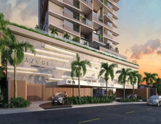 Esta é uma representação digital (renderização 3D) da fachada de um edifício residencial moderno e de vários andares, visto da rua ao nível do solo, provavelmente ao entardecer ou amanhecer. O céu ao fundo exibe tons quentes de laranja, rosa e roxo, misturados com um azul claro, característicos de um pôr do sol ou nascer do sol, com algumas nuvens suaves. O edifício em si tem uma arquitetura contemporânea. A torre principal, que se eleva acima de uma base mais larga (pódio), é predominantemente de cor clara, um tom neutro entre bege e cinza. Cada andar possui amplas varandas com guarda-corpos de vidro transparente, permitindo visibilidade e conferindo leveza ao design. Algumas varandas mostram vegetação (plantas em vasos), sugerindo espaços privativos ao ar livre para os moradores. As janelas são grandes, provavelmente portas de correr de vidro que dão acesso às varandas. A base do edifício (pódio), que ocupa os primeiros níveis, tem um design distinto. Sua fachada é revestida por um material que parece ser concreto aparente ou pedra clara, com longas faixas horizontais salientes em um tom ligeiramente mais claro, criando um padrão texturizado e rítmico. No topo desta base, há uma área com vegetação e um guarda-corpo de vidro, indicando um possível terraço ou área de lazer comum. Na fachada desta base, o nome do empreendimento "AZURE" está aplicado em letras grandes, tridimensionais e de cor escura (quase preta). Abaixo de "AZURE", lê-se "COMPACT LIFE" em letras menores, no mesmo estilo. Mais à direita, acima de uma entrada coberta, há outra placa com o que parece ser o logo "OM City". Ao nível da rua, há uma entrada para veículos delimitada por um portão moderno feito de ripas verticais de madeira (ou material que imita madeira). Um carro SUV de cor escura (parece ser um Nissan Kicks) está parado na rampa de acesso, prestes a entrar ou sair. O paisagismo é um elemento forte, com várias palmeiras altas e esguias plantadas na calçada e possivelmente dentro do terreno. Há também arbustos e vegetação rasteira verdejante ao longo da base do edifício e perto da entrada. A rua em primeiro plano é asfaltada, com faixas de trânsito amarelas pintadas. Um segundo carro, um sedan prateado, é visto em movimento na rua, ligeiramente desfocado para indicar velocidade. À direita, ao fundo, podem ser vistas mais árvores e a sugestão de outras construções de menor porte. A imagem como um todo transmite uma sensação de modernidade, sofisticação e conforto, destacando a integração do design arquitetônico com o paisagismo tropical. O empreendimento anunciado é o Apartamento com 2 Quartos a Venda Azure Compact Life Marista.
