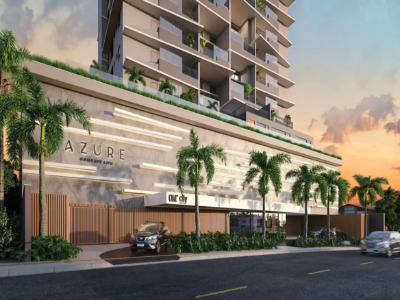 Esta é uma representação digital (renderização 3D) da fachada de um edifício residencial moderno e de vários andares, visto da rua ao nível do solo, provavelmente ao entardecer ou amanhecer. O céu ao fundo exibe tons quentes de laranja, rosa e roxo, misturados com um azul claro, característicos de um pôr do sol ou nascer do sol, com algumas nuvens suaves. O edifício em si tem uma arquitetura contemporânea. A torre principal, que se eleva acima de uma base mais larga (pódio), é predominantemente de cor clara, um tom neutro entre bege e cinza. Cada andar possui amplas varandas com guarda-corpos de vidro transparente, permitindo visibilidade e conferindo leveza ao design. Algumas varandas mostram vegetação (plantas em vasos), sugerindo espaços privativos ao ar livre para os moradores. As janelas são grandes, provavelmente portas de correr de vidro que dão acesso às varandas. A base do edifício (pódio), que ocupa os primeiros níveis, tem um design distinto. Sua fachada é revestida por um material que parece ser concreto aparente ou pedra clara, com longas faixas horizontais salientes em um tom ligeiramente mais claro, criando um padrão texturizado e rítmico. No topo desta base, há uma área com vegetação e um guarda-corpo de vidro, indicando um possível terraço ou área de lazer comum. Na fachada desta base, o nome do empreendimento "AZURE" está aplicado em letras grandes, tridimensionais e de cor escura (quase preta). Abaixo de "AZURE", lê-se "COMPACT LIFE" em letras menores, no mesmo estilo. Mais à direita, acima de uma entrada coberta, há outra placa com o que parece ser o logo "OM City". Ao nível da rua, há uma entrada para veículos delimitada por um portão moderno feito de ripas verticais de madeira (ou material que imita madeira). Um carro SUV de cor escura (parece ser um Nissan Kicks) está parado na rampa de acesso, prestes a entrar ou sair. O paisagismo é um elemento forte, com várias palmeiras altas e esguias plantadas na calçada e possivelmente dentro do terreno. Há também arbustos e vegetação rasteira verdejante ao longo da base do edifício e perto da entrada. A rua em primeiro plano é asfaltada, com faixas de trânsito amarelas pintadas. Um segundo carro, um sedan prateado, é visto em movimento na rua, ligeiramente desfocado para indicar velocidade. À direita, ao fundo, podem ser vistas mais árvores e a sugestão de outras construções de menor porte. A imagem como um todo transmite uma sensação de modernidade, sofisticação e conforto, destacando a integração do design arquitetônico com o paisagismo tropical. O empreendimento anunciado é o Apartamento com 2 Quartos a Venda Azure Compact Life Marista.