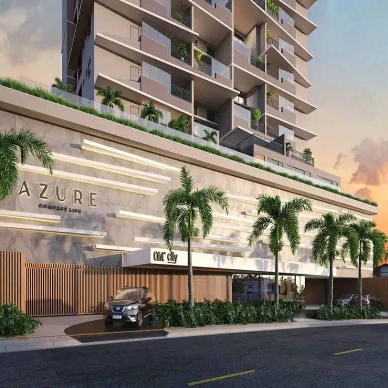 Esta é uma representação digital (renderização 3D) da fachada de um edifício residencial moderno e de vários andares, visto da rua ao nível do solo, provavelmente ao entardecer ou amanhecer. O céu ao fundo exibe tons quentes de laranja, rosa e roxo, misturados com um azul claro, característicos de um pôr do sol ou nascer do sol, com algumas nuvens suaves. O edifício em si tem uma arquitetura contemporânea. A torre principal, que se eleva acima de uma base mais larga (pódio), é predominantemente de cor clara, um tom neutro entre bege e cinza. Cada andar possui amplas varandas com guarda-corpos de vidro transparente, permitindo visibilidade e conferindo leveza ao design. Algumas varandas mostram vegetação (plantas em vasos), sugerindo espaços privativos ao ar livre para os moradores. As janelas são grandes, provavelmente portas de correr de vidro que dão acesso às varandas. A base do edifício (pódio), que ocupa os primeiros níveis, tem um design distinto. Sua fachada é revestida por um material que parece ser concreto aparente ou pedra clara, com longas faixas horizontais salientes em um tom ligeiramente mais claro, criando um padrão texturizado e rítmico. No topo desta base, há uma área com vegetação e um guarda-corpo de vidro, indicando um possível terraço ou área de lazer comum. Na fachada desta base, o nome do empreendimento "AZURE" está aplicado em letras grandes, tridimensionais e de cor escura (quase preta). Abaixo de "AZURE", lê-se "COMPACT LIFE" em letras menores, no mesmo estilo. Mais à direita, acima de uma entrada coberta, há outra placa com o que parece ser o logo "OM City". Ao nível da rua, há uma entrada para veículos delimitada por um portão moderno feito de ripas verticais de madeira (ou material que imita madeira). Um carro SUV de cor escura (parece ser um Nissan Kicks) está parado na rampa de acesso, prestes a entrar ou sair. O paisagismo é um elemento forte, com várias palmeiras altas e esguias plantadas na calçada e possivelmente dentro do terreno. Há também arbustos e vegetação rasteira verdejante ao longo da base do edifício e perto da entrada. A rua em primeiro plano é asfaltada, com faixas de trânsito amarelas pintadas. Um segundo carro, um sedan prateado, é visto em movimento na rua, ligeiramente desfocado para indicar velocidade. À direita, ao fundo, podem ser vistas mais árvores e a sugestão de outras construções de menor porte. A imagem como um todo transmite uma sensação de modernidade, sofisticação e conforto, destacando a integração do design arquitetônico com o paisagismo tropical. O empreendimento anunciado é o Apartamento com 2 Quartos a Venda Azure Compact Life Marista.
