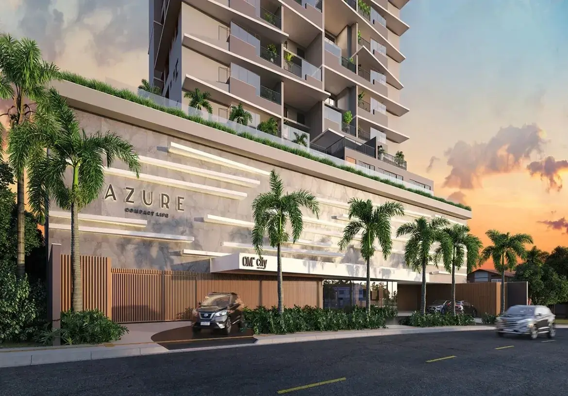 Esta é uma representação digital (renderização 3D) da fachada de um edifício residencial moderno e de vários andares, visto da rua ao nível do solo, provavelmente ao entardecer ou amanhecer. O céu ao fundo exibe tons quentes de laranja, rosa e roxo, misturados com um azul claro, característicos de um pôr do sol ou nascer do sol, com algumas nuvens suaves. O edifício em si tem uma arquitetura contemporânea. A torre principal, que se eleva acima de uma base mais larga (pódio), é predominantemente de cor clara, um tom neutro entre bege e cinza. Cada andar possui amplas varandas com guarda-corpos de vidro transparente, permitindo visibilidade e conferindo leveza ao design. Algumas varandas mostram vegetação (plantas em vasos), sugerindo espaços privativos ao ar livre para os moradores. As janelas são grandes, provavelmente portas de correr de vidro que dão acesso às varandas. A base do edifício (pódio), que ocupa os primeiros níveis, tem um design distinto. Sua fachada é revestida por um material que parece ser concreto aparente ou pedra clara, com longas faixas horizontais salientes em um tom ligeiramente mais claro, criando um padrão texturizado e rítmico. No topo desta base, há uma área com vegetação e um guarda-corpo de vidro, indicando um possível terraço ou área de lazer comum. Na fachada desta base, o nome do empreendimento "AZURE" está aplicado em letras grandes, tridimensionais e de cor escura (quase preta). Abaixo de "AZURE", lê-se "COMPACT LIFE" em letras menores, no mesmo estilo. Mais à direita, acima de uma entrada coberta, há outra placa com o que parece ser o logo "OM City". Ao nível da rua, há uma entrada para veículos delimitada por um portão moderno feito de ripas verticais de madeira (ou material que imita madeira). Um carro SUV de cor escura (parece ser um Nissan Kicks) está parado na rampa de acesso, prestes a entrar ou sair. O paisagismo é um elemento forte, com várias palmeiras altas e esguias plantadas na calçada e possivelmente dentro do terreno. Há também arbustos e vegetação rasteira verdejante ao longo da base do edifício e perto da entrada. A rua em primeiro plano é asfaltada, com faixas de trânsito amarelas pintadas. Um segundo carro, um sedan prateado, é visto em movimento na rua, ligeiramente desfocado para indicar velocidade. À direita, ao fundo, podem ser vistas mais árvores e a sugestão de outras construções de menor porte. A imagem como um todo transmite uma sensação de modernidade, sofisticação e conforto, destacando a integração do design arquitetônico com o paisagismo tropical. O empreendimento anunciado é o Apartamento com 2 Quartos a Venda Azure Compact Life Marista.