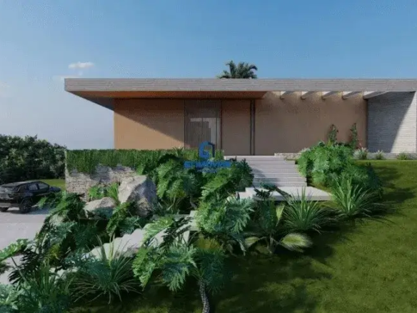 A imagem exibe uma residência de arquitetura contemporânea e linhas puras, caracterizada por um design horizontalizado que se assenta com elegância sobre um terreno em aclive. O céu de um azul intenso e sem nuvens sugere um dia de luminosidade plena, realçando cada detalhe da construção e do paisagismo. Detalhes Estruturais e Revestimentos do Imóvel: A fachada principal da casa é dominada por uma grande laje de cobertura plana que se projeta audaciosamente para a frente, formando uma generosa marquise. Esta marquise, revestida em um tom de cinza muito claro ou possivelmente um acabamento que imita madeira clara, cria uma área de sombra e transição para a entrada. Sustentando visualmente essa projeção, percebem-se pilares delgados ou extensões da parede principal, pintados no mesmo tom da fachada. A parede principal sob a marquise apresenta um revestimento liso em um tom bege suave, quase creme, conferindo uma base neutra e sofisticada. À direita da entrada, uma parede lateral se destaca por um revestimento texturizado em réguas horizontais de cor cinza claro, com variações sutis de tonalidade, lembrando pedras naturais finas como canjiquinha ou um concreto aparente trabalhado. Este contraste de texturas e cores adiciona profundidade e interesse visual à composição. A entrada principal é acessada por uma imponente escadaria de concreto claro, com degraus largos e baixos que convidam ao acesso. A porta de entrada, alta e centralizada, é um elemento de destaque, parecendo ser constituída por um grande painel de vidro com esquadrias metálicas escuras ou, alternativamente, um painel ripado vertical de madeira escura, conferindo um toque de modernidade e privacidade. Ambiente, Paisagismo e Elementos Externos: O ambiente é de exclusividade e integração com a natureza. O terreno em aclive é habilmente trabalhado com um paisagismo exuberante. Um vasto gramado de um verde vibrante e meticulosamente aparado cobre a maior parte da área frontal e lateral, ascendendo em direção à edificação. O projeto paisagístico é rico em espécies de folhagens densas e variadas. Na base da casa e margeando a escadaria, observam-se arranjos de plantas tropicais, como filodendros de folhas largas e recortadas, pequenas palmeiras ornamentais e arbustos volumosos de diferentes tonalidades de verde. Muros de contenção baixos, construídos com pedras naturais rústicas de coloração clara (bege-acinzentado), acompanham o desnível do terreno. Sobre estes muros, vegetação pendente e rasteira se derrama, suavizando as linhas e integrando as estruturas de pedra ao verde circundante. À esquerda da imagem, uma rampa de acesso para veículos, pavimentada em concreto claro e liso, desce em direção à parte mais baixa do terreno. Um veículo SUV de porte médio a grande, na cor preta ou cinza chumbo, está estacionado ao final desta rampa, sugerindo a entrada da garagem ou uma área de estacionamento. Ao fundo, delimitando visualmente a propriedade, uma densa massa de árvores altas com copas frondosas forma um pano de fundo verde escuro, garantindo privacidade e reforçando a sensação de imersão na natureza. Nota-se, à extrema direita e mais distante, o tronco nu de uma árvore, talvez uma espécie caducifólia. Cores Predominantes: A paleta de cores é sofisticada e harmoniosa: Construção: Bege claro/creme nas paredes principais, cinza claro na marquise e na parede texturizada. Paisagismo: Múltiplos tons de verde (do claro ao escuro), marrons e cinzas das pedras e troncos. Acessos: Cinza claro do concreto. Céu: Azul intenso. Móveis: Nesta perspectiva da fachada e do jardim frontal, não são visíveis quaisquer peças de mobiliário externo, como mesas, cadeiras ou espreguiçadeiras. O foco está inteiramente na arquitetura imponente do imóvel e na sua integração com o paisagismo cuidadosamente planejado. Em suma, a imagem retrata uma propriedade de alto padrão no Residencial Aldeia do Vale, onde a arquitetura moderna se funde com um paisagismo tropical exuberante, transmitindo uma atmosfera de luxo, tranquilidade e contato íntimo com a natureza.