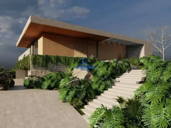 Esta imagem retrata a fachada de um imóvel de arquitetura moderna e imponente, possivelmente uma representação digital (render). Detalhes do Imóvel: Arquitetura e Estrutura: O imóvel possui um design contemporâneo, caracterizado por linhas retas e volumes bem definidos. O telhado é plano e extenso, com uma laje de cor clara (provavelmente concreto ou revestimento similar) que se projeta significativamente sobre a estrutura, criando beirais amplos. A face inferior desses beirais é revestida com um material que lembra madeira de tom quente, alaranjado/caramelo, adicionando um toque de aconchego. As paredes do nível superior parecem ser de alvenaria com acabamento liso em tom bege claro ou off-white. A base do imóvel, ou um muro de contenção/elemento paisagístico proeminente, é construído com pedras naturais de formatos irregulares, em tons de cinza, bege e marrom, conferindo um aspecto rústico e elegante que contrasta com a modernidade do restante da estrutura. Há grandes vãos e aberturas, sugerindo amplas janelas ou portas de vidro, embora o interior não seja visível. Cores Predominantes: Neutras: Bege claro/off-white (paredes superiores, laje do telhado, escadaria, piso da entrada). Naturais: Tons de cinza, bege e marrom (pedras do muro), verde intenso (vegetação), marrom/alaranjado (revestimento inferior do beiral). Escuras: Preto ou cinza escuro (SUV). Ambiente e Paisagismo: O ambiente transmite sofisticação e uma forte integração com a natureza. O paisagismo é exuberante, com destaque para plantas tropicais de folhas largas e volumosas (como filodendros ou guaimbês) em tons de verde vibrante, que margeiam a escadaria e se espalham pelo terreno. Há uma escadaria larga, feita de degraus claros (possivelmente concreto ou pedra), que leva da área de acesso de veículos ao nível principal da casa. Trepadeiras verdes cobrem parte do muro de pedras, integrando-o ainda mais ao paisagismo. No lado esquerdo, uma rampa de acesso para veículos, pavimentada com blocos retangulares de cor clara (similar à escadaria), leva a uma área onde um SUV de cor escura (preto ou cinza chumbo) está estacionado. Ao fundo e nas laterais, observa-se vegetação mais densa e um gramado bem cuidado. Uma árvore de galhos nus se destaca à direita, contrastando com a folhagem densa ao redor. O céu é parcialmente nublado, com nuvens escuras à esquerda e áreas mais claras com um pouco de azul à direita, sugerindo um final de tarde ou um dia com tempo instável, o que confere um certo drama à cena. Móveis (Elementos Externos Fixos): Não há móveis externos visíveis como cadeiras ou mesas. Os "móveis" aqui seriam os elementos construtivos e paisagísticos que compõem a cena externa. A escadaria e a rampa de acesso são elementos funcionais e estéticos importantes. Grandes rochas naturais estão integradas ao paisagismo próximo à escada, servindo como elementos decorativos. Em resumo, a imagem apresenta um imóvel de luxo com design moderno, caracterizado pelo uso de materiais naturais como pedra e madeira em contraste com superfícies lisas e claras, inserido em um ambiente com paisagismo tropical rico e bem planejado.