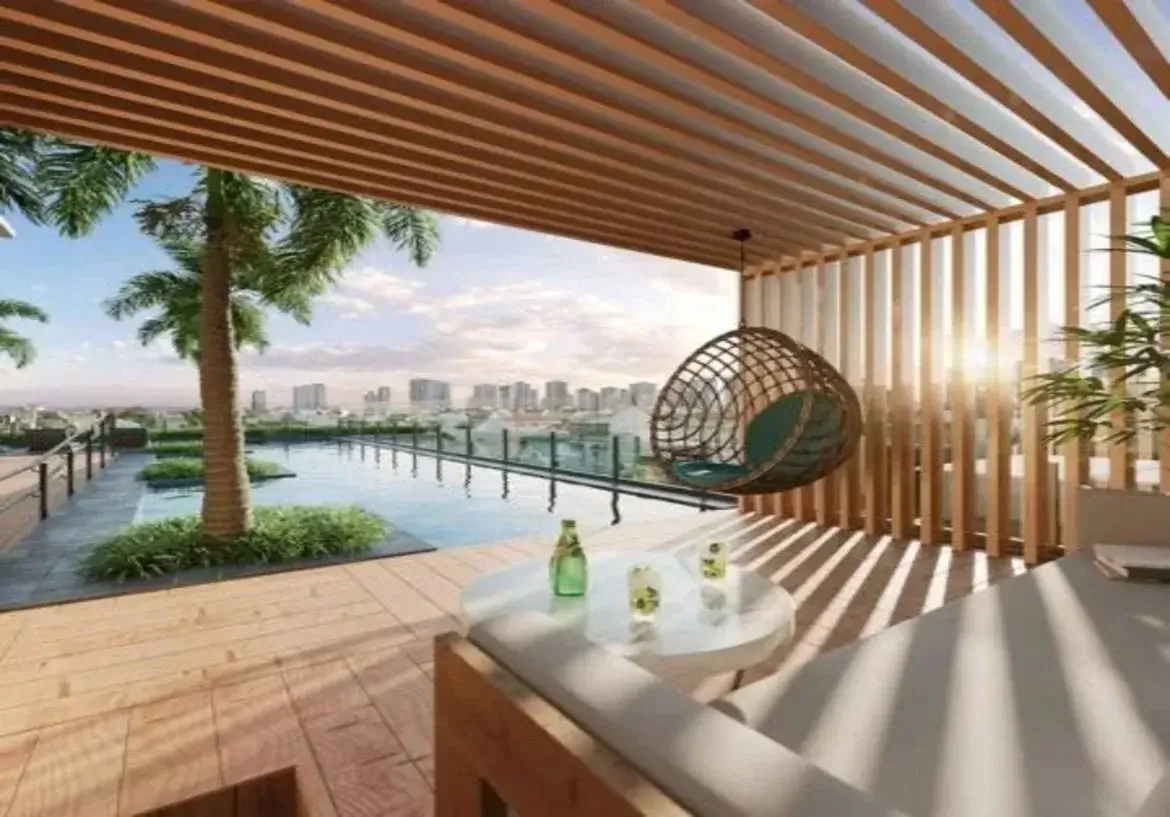 A imagem exibe uma luxuosa área externa de um imóvel, provavelmente um terraço ou varanda gourmet de um apartamento de alto padrão, com vista panorâmica para a cidade. O ambiente combina design moderno com elementos naturais, criando uma atmosfera sofisticada e relaxante. Estrutura e Cores: Cobertura e Parede Lateral: O espaço é parcialmente coberto por um pergolado de madeira clara com ripas horizontais, que se estende do teto. Uma das paredes laterais segue o mesmo padrão, com ripas de madeira verticais, também em tom claro. Essa estrutura ripada permite a passagem da luz do sol, criando um interessante jogo de luz e sombra lineares sobre o piso e os móveis, especialmente acentuado pela luz dourada do fim de tarde que entra pela lateral. Piso: O piso da área coberta é de madeira clara, em réguas, harmonizando com a estrutura do pergolado e da parede. Fora da área coberta, junto à piscina, o piso parece ser de um material diferente, talvez um deck ou piso cimentício em tom de cinza escuro. Cores Predominantes: A paleta de cores é dominada por tons neutros e naturais: madeira clara, branco/bege dos estofados, cinza escuro do piso externo, o azul da piscina e do céu, e o verde da vegetação. Há um toque de cor mais vibrante no verde-petróleo da almofada da cadeira suspensa. Ambiente e Mobiliário: Área de Estar: Sob o pergolado, há um confortável sofá ou daybed com estrutura de madeira clara e grandes almofadas/estofados em tecido de cor clara (branco ou bege). À frente do sofá, uma mesinha de centro redonda e baixa, com tampo branco ou muito claro, apoia uma garrafa verde e dois copos com bebida, sugerindo um espaço de convívio e relaxamento. Cadeira Suspensa: Um destaque do mobiliário é uma cadeira suspensa estilo "ovo" ou "ninho", feita de vime ou material sintético trançado em tom marrom escuro. Ela possui uma almofada interna em um tom de verde-petróleo ou azul-esverdeado escuro e está pendurada na estrutura do pergolado. Área da Piscina: Adjacente à área coberta, há uma piscina de borda infinita com água azul cristalina. A borda da piscina é protegida por um guarda-corpo de vidro transparente com suportes discretos, garantindo segurança sem obstruir a vista. Paisagismo: Palmeiras altas e vegetação rasteira verde estão plantadas junto à piscina, trazendo um elemento tropical e natural para o ambiente urbano. Uma planta em vaso também é visível no canto da área coberta. Vista e Iluminação: Vista: O imóvel oferece uma vista deslumbrante e ampla da cidade, com um skyline repleto de edifícios ao fundo. Iluminação: A cena é banhada pela luz natural do sol, que parece estar baixo no horizonte (fim de tarde/pôr do sol), criando reflexos na piscina, sombras alongadas e uma atmosfera quente e convidativa. Em resumo, a imagem retrata um espaço externo elegante e bem projetado, ideal para lazer e contemplação, que valoriza a vista urbana e utiliza materiais naturais e design contemporâneo para criar um refúgio sofisticado e acolhedor.
