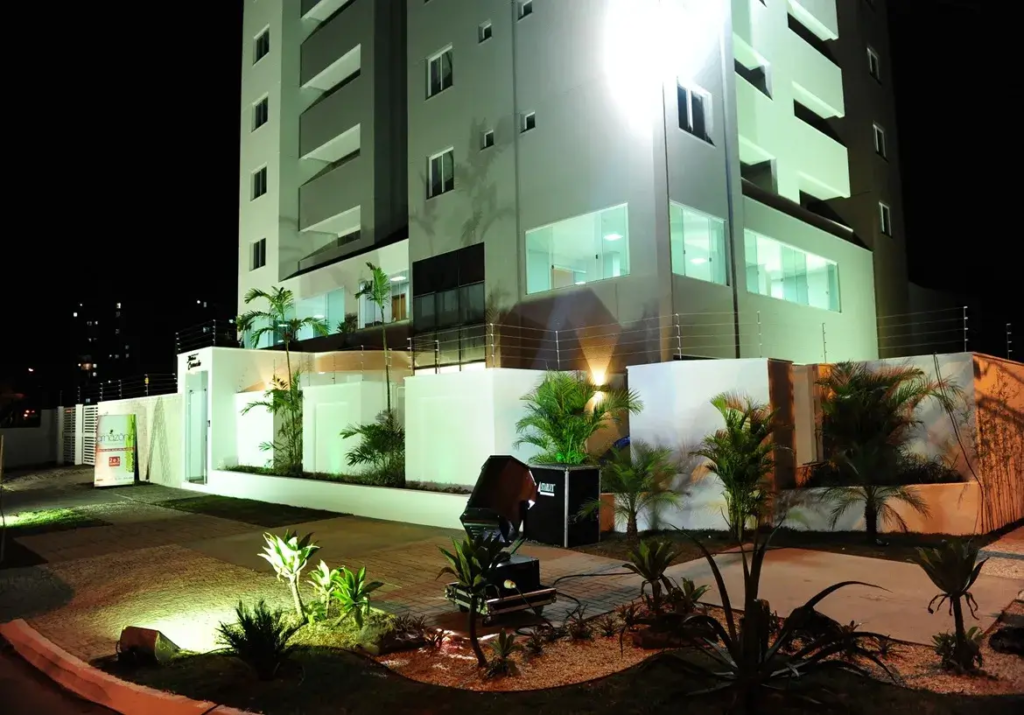 Esta imagem noturna captura a fachada imponente e moderna do Residencial Flor do Cerrado.O edifício principal, com múltiplos andares, exibe uma arquitetura contemporânea, com paredes predominantemente em um tom claro, possivelmente branco ou um cinza muito pálido, que é intensamente iluminado por um potente refletor, conferindo-lhe um brilho destacado contra o céu escuro. As sacadas são embutidas e possuem um tom de cinza um pouco mais escuro, criando um contraste sutil e elegante. Janelas de vidro refletem a iluminação, e algumas luzes internas acesas sugerem atividade ou apartamentos modelo.No térreo e na área externa, o ambiente é cuidadosamente planejado e bem iluminado. Muros brancos delimitam a propriedade, coroados por uma cerca elétrica visível, indicando preocupação com a segurança. A entrada parece ser marcada por uma estrutura também branca à esquerda, onde se avista um banner ou placa informativa, parcialmente visível.O paisagismo é um destaque, com diversas plantas ornamentais, incluindo palmeiras de pequeno e médio porte e arbustos, dispostas em canteiros com cobertura de pedrisco claro. A iluminação do jardim é feita por spots direcionados, que realçam a vegetação e criam um jogo de luz e sombra agradável. Um caminho pavimentado, possivelmente com blocos intertravados, conduz pela área externa.Um equipamento de iluminação profissional, um grande refletor preto sobre um suporte, está posicionado em primeiro plano no jardim, direcionado para o edifício, o que sugere um evento de lançamento ou uma iluminação especial para destacar o empreendimento. Não há móveis externos visíveis, exceto por este equipamento de iluminação.O ambiente geral é sofisticado e acolhedor, transmitindo uma sensação de modernidade, segurança e cuidado com os detalhes, característicos de um empreendimento novo e de qualidade como o Residencial Flor do Cerrado.