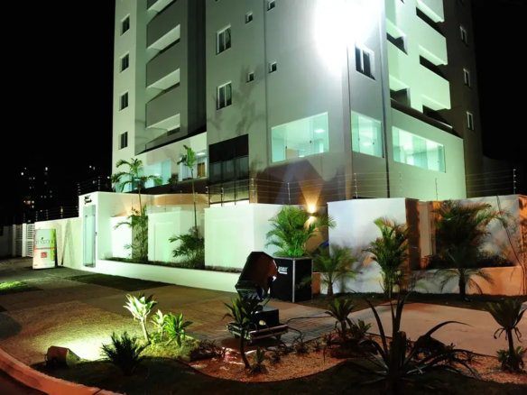 residencial-flor-do-cerrado-fachada Esta imagem noturna captura a fachada imponente e moderna do Residencial Flor do Cerrado. O edifício principal, com múltiplos andares, exibe uma arquitetura contemporânea, com paredes predominantemente em um tom claro, possivelmente branco ou um cinza muito pálido, que é intensamente iluminado por um potente refletor, conferindo-lhe um brilho destacado contra o céu escuro. As sacadas são embutidas e possuem um tom de cinza um pouco mais escuro, criando um contraste sutil e elegante. Janelas de vidro refletem a iluminação, e algumas luzes internas acesas sugerem atividade ou apartamentos modelo. No térreo e na área externa, o ambiente é cuidadosamente planejado e bem iluminado. Muros brancos delimitam a propriedade, coroados por uma cerca elétrica visível, indicando preocupação com a segurança. A entrada parece ser marcada por uma estrutura também branca à esquerda, onde se avista um banner ou placa informativa, parcialmente visível. O paisagismo é um destaque, com diversas plantas ornamentais, incluindo palmeiras de pequeno e médio porte e arbustos, dispostas em canteiros com cobertura de pedrisco claro. A iluminação do jardim é feita por spots direcionados, que realçam a vegetação e criam um jogo de luz e sombra agradável. Um caminho pavimentado, possivelmente com blocos intertravados, conduz pela área externa. Um equipamento de iluminação profissional, um grande refletor preto sobre um suporte, está posicionado em primeiro plano no jardim, direcionado para o edifício, o que sugere um evento de lançamento ou uma iluminação especial para destacar o empreendimento. Não há móveis externos visíveis, exceto por este equipamento de iluminação. O ambiente geral é sofisticado e acolhedor, transmitindo uma sensação de modernidade, segurança e cuidado com os detalhes, característicos de um empreendimento novo e de qualidade como o Residencial Flor do Cerrado.