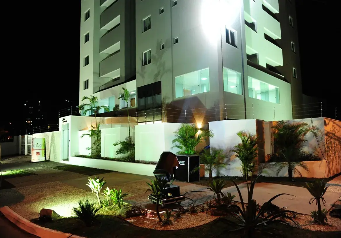 residencial-flor-do-cerrado-fachada Esta imagem noturna captura a fachada imponente e moderna do Residencial Flor do Cerrado. O edifício principal, com múltiplos andares, exibe uma arquitetura contemporânea, com paredes predominantemente em um tom claro, possivelmente branco ou um cinza muito pálido, que é intensamente iluminado por um potente refletor, conferindo-lhe um brilho destacado contra o céu escuro. As sacadas são embutidas e possuem um tom de cinza um pouco mais escuro, criando um contraste sutil e elegante. Janelas de vidro refletem a iluminação, e algumas luzes internas acesas sugerem atividade ou apartamentos modelo. No térreo e na área externa, o ambiente é cuidadosamente planejado e bem iluminado. Muros brancos delimitam a propriedade, coroados por uma cerca elétrica visível, indicando preocupação com a segurança. A entrada parece ser marcada por uma estrutura também branca à esquerda, onde se avista um banner ou placa informativa, parcialmente visível. O paisagismo é um destaque, com diversas plantas ornamentais, incluindo palmeiras de pequeno e médio porte e arbustos, dispostas em canteiros com cobertura de pedrisco claro. A iluminação do jardim é feita por spots direcionados, que realçam a vegetação e criam um jogo de luz e sombra agradável. Um caminho pavimentado, possivelmente com blocos intertravados, conduz pela área externa. Um equipamento de iluminação profissional, um grande refletor preto sobre um suporte, está posicionado em primeiro plano no jardim, direcionado para o edifício, o que sugere um evento de lançamento ou uma iluminação especial para destacar o empreendimento. Não há móveis externos visíveis, exceto por este equipamento de iluminação. O ambiente geral é sofisticado e acolhedor, transmitindo uma sensação de modernidade, segurança e cuidado com os detalhes, característicos de um empreendimento novo e de qualidade como o Residencial Flor do Cerrado.