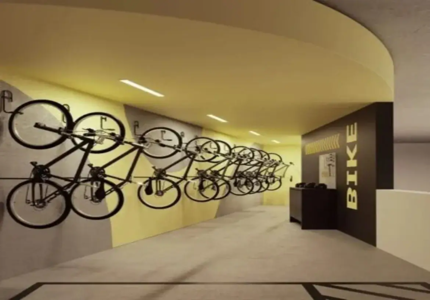 Esta imagem retrata o bicicletário moderno e bem organizado do edifício residencial Azure Compact Life. Ambiente e Cores: O ambiente é um corredor, com uma perspectiva que se aprofunda na imagem. A iluminação é feita por luminárias lineares embutidas no teto, que fornecem uma luz clara e uniforme. A paleta de cores é marcante e contemporânea: Teto: Um teto rebaixado e curvo na entrada é pintado em um tom vibrante de amarelo mostarda, que se estende por todo o comprimento visível do corredor. Paredes: A parede principal à esquerda é predominantemente cinza claro, mas possui um grafismo dinâmico com uma faixa diagonal ascendente em amarelo (mesmo tom do teto) que ocupa a parte superior. Ao fundo à direita, uma parede se destaca em um tom escuro, provavelmente preto ou marrom bem escuro, servindo como pano de fundo para sinalização. Uma parede branca é visível mais ao fundo à direita, criando contraste. Piso: O piso é de concreto liso ou um material similar em tom de cinza claro, com faixas pretas demarcando áreas ou caminhos. Móveis e Detalhes: Bicicletas e Suportes: O principal elemento são as diversas bicicletas pretas, todas aparentemente do mesmo modelo (estilo urbano ou passeio), meticulosamente dispostas ao longo da parede esquerda. Elas estão fixadas em suportes metálicos individuais, organizadas em duas alturas alternadas para otimizar o espaço: uma fileira mais alta, pendurada pela roda dianteira e apoiada no pedal, e uma fileira mais baixa, também suspensa, mas posicionada abaixo da primeira. Sinalização: Na parede escura à direita, a palavra "BIKE" está escrita em letras maiúsculas amarelas, na vertical, acompanhada por setas amarelas (chevrons) apontando para a esquerda e um pequeno quadro ou cartaz, sugerindo a direção ou a identificação da área. Móvel de Apoio: Próximo a essa parede escura, há um móvel baixo e retangular, também na cor preta ou marrom escura, que pode servir como bancada de apoio, para guardar capacetes, ferramentas ou outros acessórios, embora não haja objetos visíveis sobre ele. O design geral é funcional e estilizado, utilizando cores contrastantes e elementos gráficos para criar um espaço prático e visualmente interessante para o armazenamento de bicicletas. A imagem retrata um bicicletário moderno e bem organizado, provavelmente localizado no interior de um edifício residencial ou comercial. Ambiente e Cores: O ambiente é um corredor, com uma perspectiva que se aprofunda na imagem. A iluminação é feita por luminárias lineares embutidas no teto, que fornecem uma luz clara e uniforme. A paleta de cores é marcante e contemporânea: Teto: Um teto rebaixado e curvo na entrada é pintado em um tom vibrante de amarelo mostarda, que se estende por todo o comprimento visível do corredor. Paredes: A parede principal à esquerda é predominantemente cinza claro, mas possui um grafismo dinâmico com uma faixa diagonal ascendente em amarelo (mesmo tom do teto) que ocupa a parte superior. Ao fundo à direita, uma parede se destaca em um tom escuro, provavelmente preto ou marrom bem escuro, servindo como pano de fundo para sinalização. Uma parede branca é visível mais ao fundo à direita, criando contraste. Piso: O piso é de concreto liso ou um material similar em tom de cinza claro, com faixas pretas demarcando áreas ou caminhos. Móveis e Detalhes: Bicicletas e Suportes: O principal elemento são as diversas bicicletas pretas, todas aparentemente do mesmo modelo (estilo urbano ou passeio), meticulosamente dispostas ao longo da parede esquerda. Elas estão fixadas em suportes metálicos individuais, organizadas em duas alturas alternadas para otimizar o espaço: uma fileira mais alta, pendurada pela roda dianteira e apoiada no pedal, e uma fileira mais baixa, também suspensa, mas posicionada abaixo da primeira. Sinalização: Na parede escura à direita, a palavra "BIKE" está escrita em letras maiúsculas amarelas, na vertical, acompanhada por setas amarelas (chevrons) apontando para a esquerda e um pequeno quadro ou cartaz, sugerindo a direção ou a identificação da área. Móvel de Apoio: Próximo a essa parede escura, há um móvel baixo e retangular, também na cor preta ou marrom escura, que pode servir como bancada de apoio, para guardar capacetes, ferramentas ou outros acessórios, embora não haja objetos visíveis sobre ele. O design geral é funcional e estilizado, utilizando cores contrastantes e elementos gráficos para criar um espaço prático e visualmente interessante para o armazenamento de