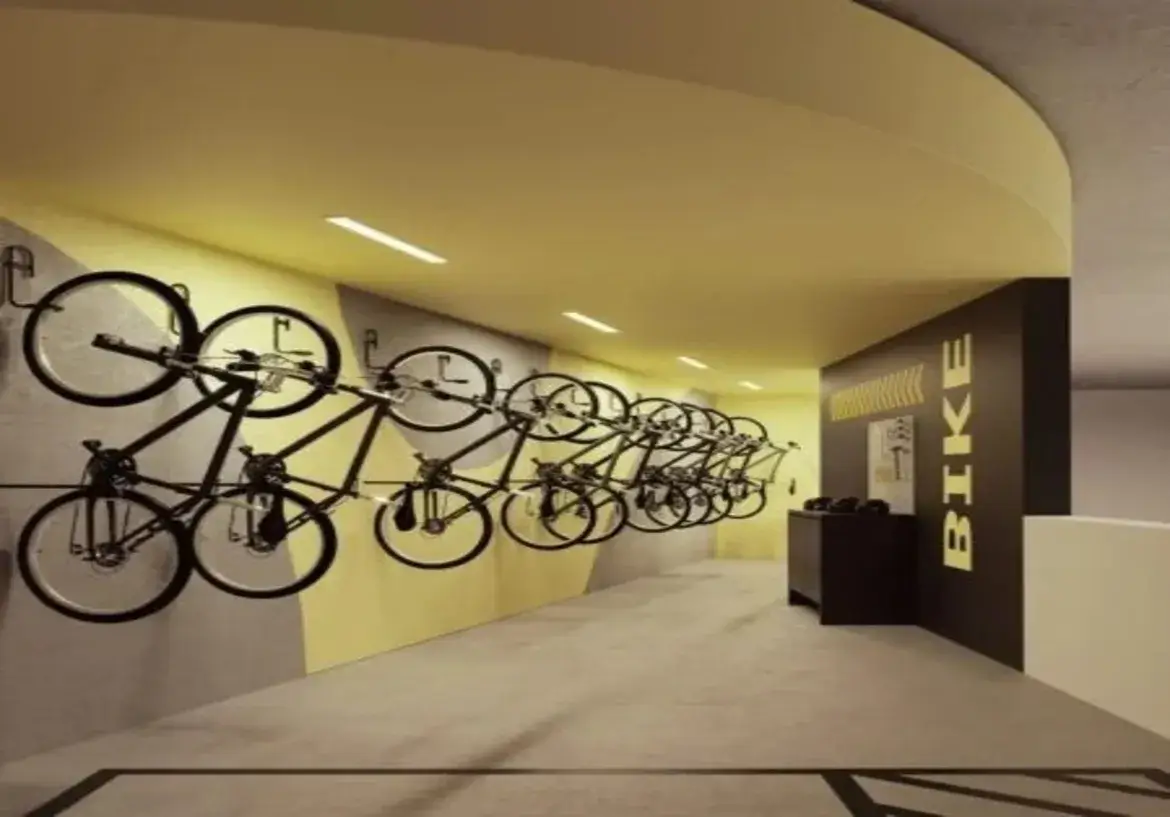 Esta imagem retrata o bicicletário moderno e bem organizado do edifício residencial Azure Compact Life. Ambiente e Cores: O ambiente é um corredor, com uma perspectiva que se aprofunda na imagem. A iluminação é feita por luminárias lineares embutidas no teto, que fornecem uma luz clara e uniforme. A paleta de cores é marcante e contemporânea: Teto: Um teto rebaixado e curvo na entrada é pintado em um tom vibrante de amarelo mostarda, que se estende por todo o comprimento visível do corredor. Paredes: A parede principal à esquerda é predominantemente cinza claro, mas possui um grafismo dinâmico com uma faixa diagonal ascendente em amarelo (mesmo tom do teto) que ocupa a parte superior. Ao fundo à direita, uma parede se destaca em um tom escuro, provavelmente preto ou marrom bem escuro, servindo como pano de fundo para sinalização. Uma parede branca é visível mais ao fundo à direita, criando contraste. Piso: O piso é de concreto liso ou um material similar em tom de cinza claro, com faixas pretas demarcando áreas ou caminhos. Móveis e Detalhes: Bicicletas e Suportes: O principal elemento são as diversas bicicletas pretas, todas aparentemente do mesmo modelo (estilo urbano ou passeio), meticulosamente dispostas ao longo da parede esquerda. Elas estão fixadas em suportes metálicos individuais, organizadas em duas alturas alternadas para otimizar o espaço: uma fileira mais alta, pendurada pela roda dianteira e apoiada no pedal, e uma fileira mais baixa, também suspensa, mas posicionada abaixo da primeira. Sinalização: Na parede escura à direita, a palavra "BIKE" está escrita em letras maiúsculas amarelas, na vertical, acompanhada por setas amarelas (chevrons) apontando para a esquerda e um pequeno quadro ou cartaz, sugerindo a direção ou a identificação da área. Móvel de Apoio: Próximo a essa parede escura, há um móvel baixo e retangular, também na cor preta ou marrom escura, que pode servir como bancada de apoio, para guardar capacetes, ferramentas ou outros acessórios, embora não haja objetos visíveis sobre ele. O design geral é funcional e estilizado, utilizando cores contrastantes e elementos gráficos para criar um espaço prático e visualmente interessante para o armazenamento de bicicletas. A imagem retrata um bicicletário moderno e bem organizado, provavelmente localizado no interior de um edifício residencial ou comercial. Ambiente e Cores: O ambiente é um corredor, com uma perspectiva que se aprofunda na imagem. A iluminação é feita por luminárias lineares embutidas no teto, que fornecem uma luz clara e uniforme. A paleta de cores é marcante e contemporânea: Teto: Um teto rebaixado e curvo na entrada é pintado em um tom vibrante de amarelo mostarda, que se estende por todo o comprimento visível do corredor. Paredes: A parede principal à esquerda é predominantemente cinza claro, mas possui um grafismo dinâmico com uma faixa diagonal ascendente em amarelo (mesmo tom do teto) que ocupa a parte superior. Ao fundo à direita, uma parede se destaca em um tom escuro, provavelmente preto ou marrom bem escuro, servindo como pano de fundo para sinalização. Uma parede branca é visível mais ao fundo à direita, criando contraste. Piso: O piso é de concreto liso ou um material similar em tom de cinza claro, com faixas pretas demarcando áreas ou caminhos. Móveis e Detalhes: Bicicletas e Suportes: O principal elemento são as diversas bicicletas pretas, todas aparentemente do mesmo modelo (estilo urbano ou passeio), meticulosamente dispostas ao longo da parede esquerda. Elas estão fixadas em suportes metálicos individuais, organizadas em duas alturas alternadas para otimizar o espaço: uma fileira mais alta, pendurada pela roda dianteira e apoiada no pedal, e uma fileira mais baixa, também suspensa, mas posicionada abaixo da primeira. Sinalização: Na parede escura à direita, a palavra "BIKE" está escrita em letras maiúsculas amarelas, na vertical, acompanhada por setas amarelas (chevrons) apontando para a esquerda e um pequeno quadro ou cartaz, sugerindo a direção ou a identificação da área. Móvel de Apoio: Próximo a essa parede escura, há um móvel baixo e retangular, também na cor preta ou marrom escura, que pode servir como bancada de apoio, para guardar capacetes, ferramentas ou outros acessórios, embora não haja objetos visíveis sobre ele. O design geral é funcional e estilizado, utilizando cores contrastantes e elementos gráficos para criar um espaço prático e visualmente interessante para o armazenamento de