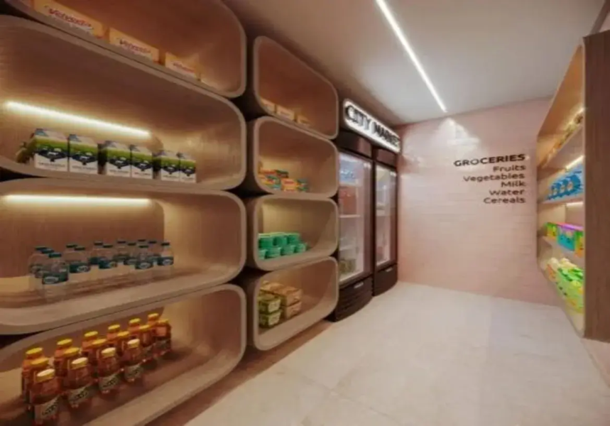 A imagem mostra o interior de um pequeno mercado ou minimercado moderno, provavelmente localizado dentro de um edifício residencial ou comercial (um "minimarket" ou "honest market"). O espaço é relativamente estreito, configurado como um corredor. Paredes e Cores: A parede esquerda é dominada por um sistema de estantes modulares de madeira clara ou laminado com tom amadeirado. A parede do fundo, onde estão as geladeiras, e a parede direita são pintadas em um tom suave de rosa claro ou terracota pálido. A parede direita parece ter uma textura sutil, talvez de tijolinhos pintados ou um revestimento similar. O teto é branco. Mobiliário e Estruturas: Estantes (Parede Esquerda): O destaque principal são as prateleiras modulares com um design único. Elas são compostas por nichos grandes e profundos com cantos arredondados, lembrando cápsulas ou retângulos suavizados, empilhados uns sobre os outros. O interior dos nichos é do mesmo material de madeira clara. Cada nicho principal possui iluminação embutida (provavelmente fita de LED) na parte superior interna, que projeta uma luz quente para baixo, iluminando os produtos. Geladeiras (Fundo): Ao fundo, há duas geladeiras expositoras verticais, com portas de vidro e molduras escuras (pretas ou marrom-escuras). Acima delas, um letreiro luminoso branco com letras escuras indica "CITY MARKET". Estantes (Parede Direita): A parede direita possui prateleiras mais simples, embutidas na parede, também em madeira clara e com iluminação embutida similar às da esquerda. Piso: O piso é revestido com ladrilhos grandes de cor clara, talvez um cinza-claro ou bege, com um acabamento liso e levemente reflexivo. Iluminação: Além da iluminação embutida nas prateleiras, há uma luminária linear longa e fina embutida no teto branco, percorrendo o comprimento do corredor e fornecendo a iluminação geral do ambiente. A luz geral parece ser branca e funcional, contrastando com a luz mais quente e âmbar das prateleiras. Conteúdo e Ambiente: As prateleiras estão abastecidas com produtos típicos de conveniência: caixas de leite ou suco, garrafas de água, outras bebidas em garrafas menores (sucos, chás), e outros itens embalados (snacks, talvez cereais ou enlatados). Na parede direita, há uma inscrição em letras pretas que diz "GROCERIES" e lista categorias como "Fruits", "Vegetables", "Milk", "Water", "Cereals", indicando a seção de produtos. O ambiente geral é limpo, organizado e moderno, com um design que mistura funcionalidade e estética. As formas arredondadas das estantes e a combinação da madeira clara com o rosa pálido criam uma atmosfera acolhedora e contemporânea, apesar do espaço reduzido. A iluminação direcionada nas prateleiras valoriza os produtos expostos.