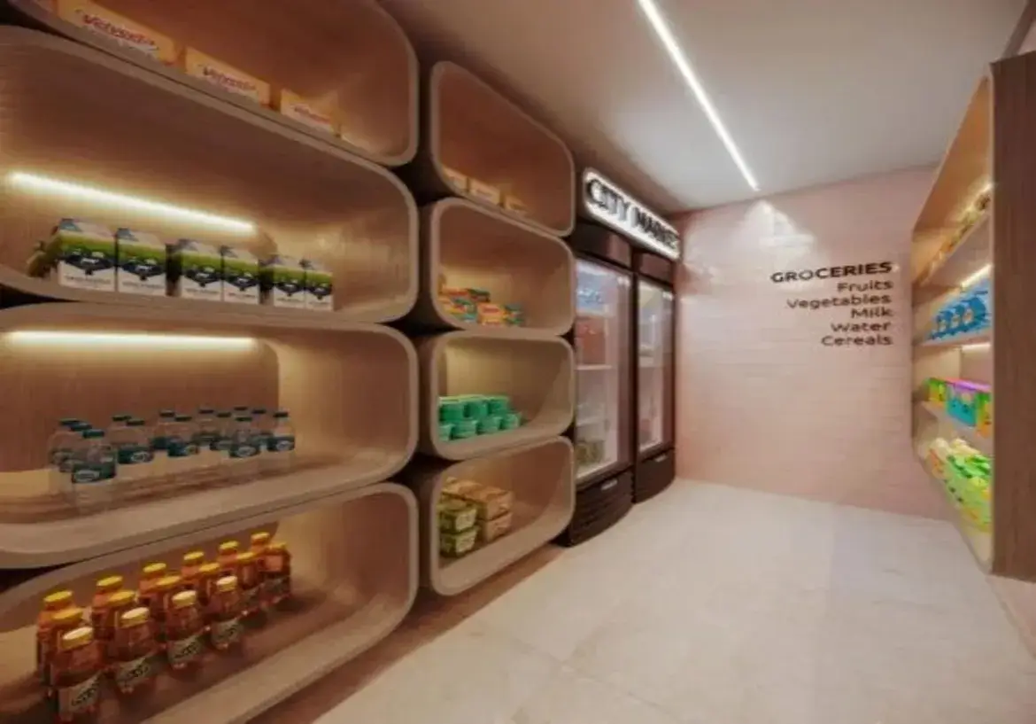 A imagem mostra o interior de um pequeno mercado ou minimercado moderno, provavelmente localizado dentro de um edifício residencial ou comercial (um "minimarket" ou "honest market"). O espaço é relativamente estreito, configurado como um corredor. Paredes e Cores: A parede esquerda é dominada por um sistema de estantes modulares de madeira clara ou laminado com tom amadeirado. A parede do fundo, onde estão as geladeiras, e a parede direita são pintadas em um tom suave de rosa claro ou terracota pálido. A parede direita parece ter uma textura sutil, talvez de tijolinhos pintados ou um revestimento similar. O teto é branco. Mobiliário e Estruturas: Estantes (Parede Esquerda): O destaque principal são as prateleiras modulares com um design único. Elas são compostas por nichos grandes e profundos com cantos arredondados, lembrando cápsulas ou retângulos suavizados, empilhados uns sobre os outros. O interior dos nichos é do mesmo material de madeira clara. Cada nicho principal possui iluminação embutida (provavelmente fita de LED) na parte superior interna, que projeta uma luz quente para baixo, iluminando os produtos. Geladeiras (Fundo): Ao fundo, há duas geladeiras expositoras verticais, com portas de vidro e molduras escuras (pretas ou marrom-escuras). Acima delas, um letreiro luminoso branco com letras escuras indica "CITY MARKET". Estantes (Parede Direita): A parede direita possui prateleiras mais simples, embutidas na parede, também em madeira clara e com iluminação embutida similar às da esquerda. Piso: O piso é revestido com ladrilhos grandes de cor clara, talvez um cinza-claro ou bege, com um acabamento liso e levemente reflexivo. Iluminação: Além da iluminação embutida nas prateleiras, há uma luminária linear longa e fina embutida no teto branco, percorrendo o comprimento do corredor e fornecendo a iluminação geral do ambiente. A luz geral parece ser branca e funcional, contrastando com a luz mais quente e âmbar das prateleiras. Conteúdo e Ambiente: As prateleiras estão abastecidas com produtos típicos de conveniência: caixas de leite ou suco, garrafas de água, outras bebidas em garrafas menores (sucos, chás), e outros itens embalados (snacks, talvez cereais ou enlatados). Na parede direita, há uma inscrição em letras pretas que diz "GROCERIES" e lista categorias como "Fruits", "Vegetables", "Milk", "Water", "Cereals", indicando a seção de produtos. O ambiente geral é limpo, organizado e moderno, com um design que mistura funcionalidade e estética. As formas arredondadas das estantes e a combinação da madeira clara com o rosa pálido criam uma atmosfera acolhedora e contemporânea, apesar do espaço reduzido. A iluminação direcionada nas prateleiras valoriza os produtos expostos.