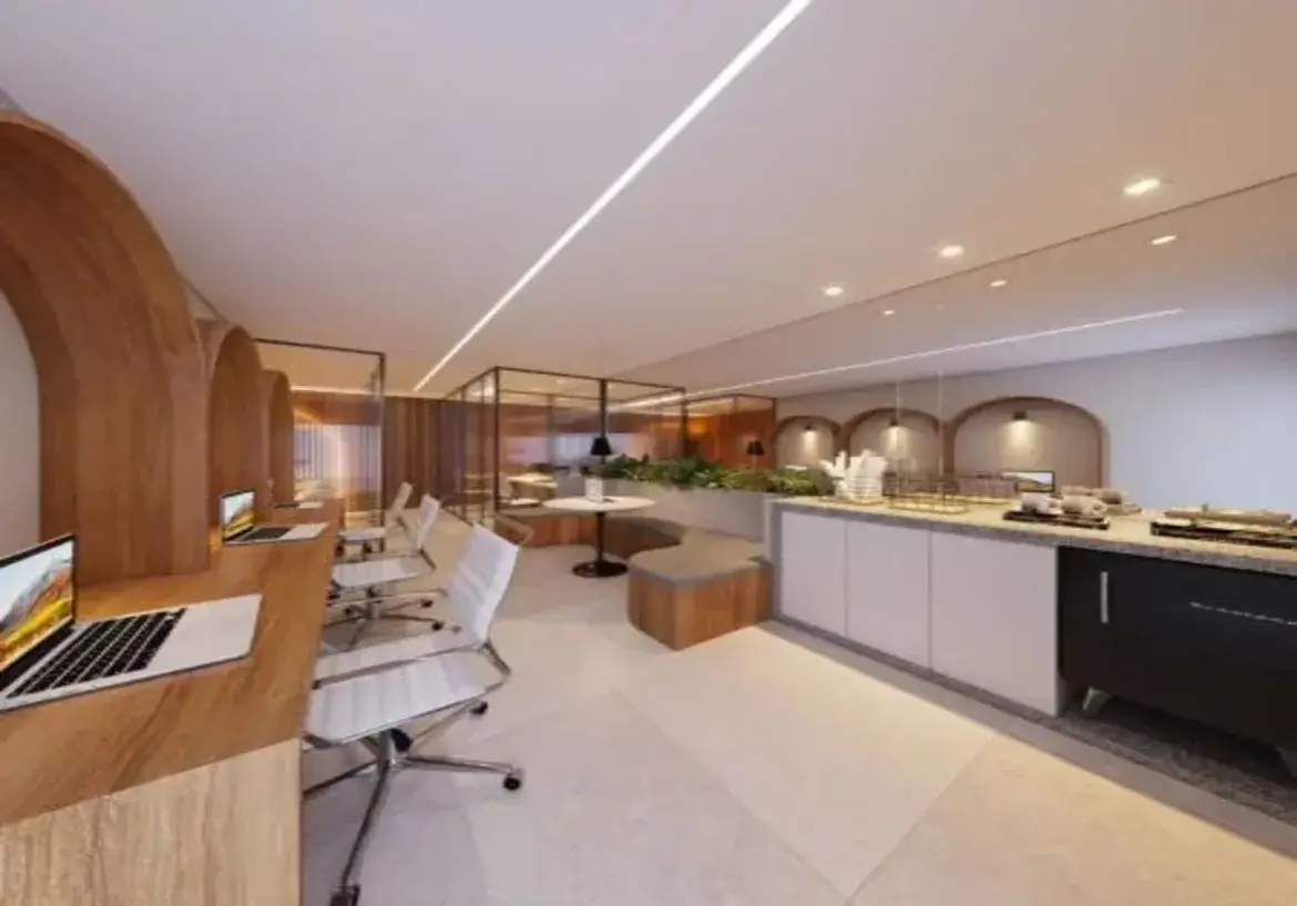 Visão Geral do Ambiente: A imagem retrata um espaço de trabalho ou convivência moderno e bem iluminado, provavelmente um coworking, lounge de escritório ou área comum de um edifício residencial/comercial. A atmosfera é sofisticada e funcional, combinando elementos de design contemporâneo com materiais quentes. Detalhes do Imóvel: Layout e Estrutura: O espaço é amplo e parece dividido em zonas funcionais distintas. À esquerda, há uma área de trabalho individualizada; no centro, uma zona de estar mais informal; e à direita, uma copa ou área de café. Ao fundo, percebe-se uma área separada por divisórias de vidro. Cores Predominantes: A paleta de cores é majoritariamente neutra e clara, contribuindo para a sensação de amplitude e luminosidade. Predominam o branco (teto, paredes, cadeiras, parte dos armários), tons de bege/creme (piso, bancada), cinza (estofado do banco, tampo da bancada) e o tom quente da madeira clara/média (arcos, mesas, base do banco, painéis). O preto surge em detalhes pontuais (bases das mesas, luminárias pendentes, molduras das divisórias de vidro, eletrodoméstico). O verde das plantas adiciona um toque de cor natural. Mobiliário e Acabamentos: Área de Trabalho (Esquerda): O destaque são as grandes estruturas em arco feitas de madeira clara, que criam estações de trabalho semi-privativas. Cada nicho possui uma bancada de trabalho do mesmo material, equipada com um laptop. As cadeiras são de escritório, modelo "Eames" ou similar, com assento e encosto brancos (provavelmente couro sintético ou plástico) e base cromada giratória com rodízios. Área de Estar (Centro): Um banco curvo embutido acompanha a parede, com base revestida em madeira e assento estofado em tecido cinza. À frente dele, uma pequena mesa redonda com tampo branco e base preta. Atrás do banco, um canteiro elevado com plantas verdes. Copa/Balcão (Direita): Uma longa bancada com tampo que parece ser de pedra (granito ou quartzo) em tons de cinza/bege salpicado. Os armários inferiores são predominantemente brancos, com puxadores discretos ou inexistentes (sistema de clique). Há um eletrodoméstico preto embutido (possivelmente um frigobar ou adega pequena). Sobre a bancada, há itens típicos de uma copa: suportes com copos, bandejas, talvez uma máquina de café (parcialmente visível). Fundo: Divisórias de vidro com esquadrias pretas separam outra área, onde se pode ver mais painéis de madeira e possivelmente mais mesas. Iluminação: O teto é branco e liso. A iluminação é um ponto forte do design: Uma longa linha de luz LED embutida atravessa o teto diagonalmente, oferecendo iluminação geral e um elemento de design marcante. Spots de luz embutidos (downlights) estão distribuídos pelo teto, principalmente sobre a área da copa. Luminárias pendentes com cúpulas pretas são usadas como iluminação de destaque sobre a mesa redonda central e dentro dos nichos decorativos na parede atrás da copa. Piso: O piso é revestido com peças grandes de porcelanato ou material similar, em tom bege claro ou off-white, de acabamento fosco ou acetinado. Paredes e Detalhes Arquitetônicos: As paredes são predominantemente brancas. Um detalhe interessante é a repetição do motivo de arco: os grandes arcos de madeira das estações de trabalho são ecoados por nichos em arco menores na parede atrás da copa, cada um com uma luminária pendente, criando ritmo e coesão visual. Ambiência Geral: O ambiente transmite uma sensação de profissionalismo e conforto. É um espaço projetado para ser ao mesmo tempo funcional para o trabalho e agradável para pausas ou interações sociais. O uso da madeira e das plantas aquece o design moderno e predominantemente neutro, tornando-o acolhedor. A iluminação bem planejada contribui para um espaço claro e convidativo.