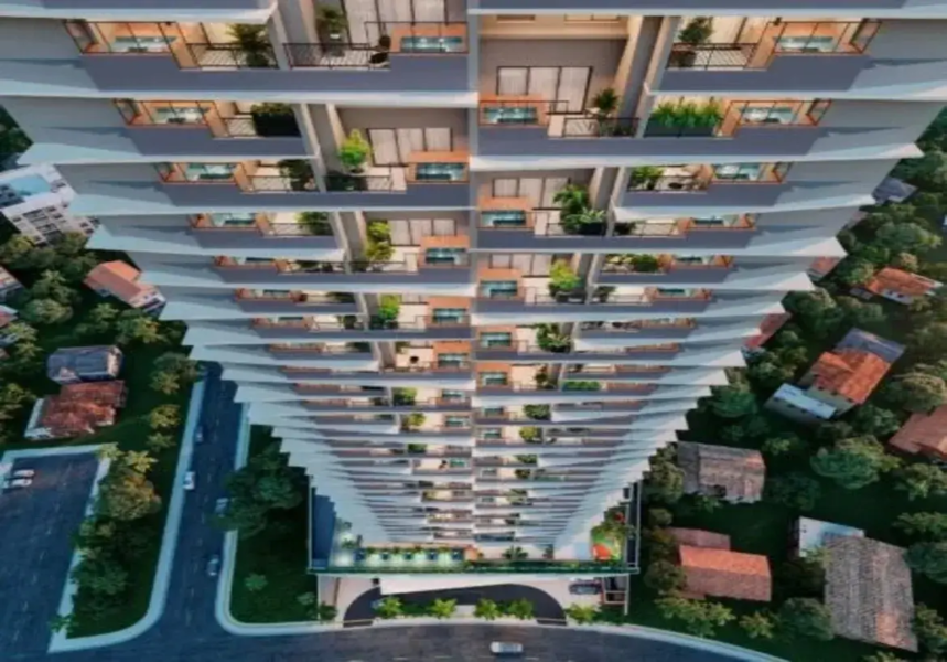 Ok, vamos descrever a imagem detalhadamente: A imagem apresenta uma vista aérea em perspectiva acentuada, olhando de cima para baixo ao longo da fachada de um edifício residencial muito alto e moderno. A perspectiva cria uma sensação de vertigem e destaca a altura imponente do prédio. Detalhes do Imóvel (Edifício): Estrutura e Design: O edifício possui múltiplos andares (um arranha-céu) com uma arquitetura contemporânea. A fachada não é reta, mas sim escalonada ou em degraus, onde cada andar ou conjunto de andares parece ligeiramente deslocado em relação ao inferior/superior, criando um efeito visual dinâmico e texturizado. Cores: A cor predominante da estrutura do edifício é um cinza claro ou branco-acinzentado. As esquadrias das grandes portas de vidro que dão para as varandas e os guarda-corpos parecem ser de um tom escuro, provavelmente preto ou cinza-chumbo. Há também detalhes que parecem ser de madeira clara ou material que imita madeira, visíveis em algumas partes das varandas (possivelmente no piso ou em painéis decorativos/divisórias). Varandas: Cada apartamento possui uma varanda individual. Elas são um elemento central do design. Guarda-corpos: Parecem ser uma combinação de metal escuro com painéis de vidro transparente ou barras verticais finas, garantindo segurança sem obstruir totalmente a vista. Piso: O piso das varandas parece ser claro, talvez cerâmico ou cimentício. Iluminação: A iluminação interna dos apartamentos é visível através das grandes portas de vidro, emitindo uma luz quente e amarelada, sugerindo que a foto foi tirada ao entardecer ou à noite. Algumas varandas também podem ter iluminação própria sutil. Ambiente e Mobiliário (Visível nas Varandas): Móveis: Muitas varandas estão decoradas com móveis de exterior. É possível distinguir pequenas mesas e cadeiras, típicas de conjuntos de bistrô ou áreas de estar compactas. A variedade indica personalização por parte dos moradores. Vegetação: Um detalhe muito relevante é a presença abundante de plantas e flores em vasos em quase todas as varandas. Há vasos de diferentes tamanhos, com plantas de folhagens verdes variadas, algumas mais altas, outras mais baixas, conferindo vida, cor e um aspecto mais natural e acolhedor à fachada predominantemente geométrica e mineral. Ambiente Geral: As luzes acesas e a presença de móveis e plantas sugerem que os apartamentos estão ocupados e as varandas são utilizadas como espaços de convivência e lazer. Entorno: Imediações: O edifício está inserido em uma área urbana, mas com bastante verde. Ao redor da base do prédio, veem-se árvores densas e outras construções mais baixas, principalmente casas com telhados de cerâmica (cor de telha / marrom-avermelhado). Há um contraste nítido entre a escala e modernidade do arranha-céu e as construções vizinhas mais tradicionais. Infraestrutura: Na parte inferior da imagem, são visíveis ruas asfaltadas com alguns carros estacionados e em movimento. Há também calçadas e o acesso à entrada do edifício, que parece ter uma área coberta (porte-cochère) e paisagismo na entrada. Em resumo, a imagem retrata um imponente edifício residencial moderno de cor clara (cinza/branco) com detalhes escuros e em madeira, caracterizado por sua fachada escalonada e varandas individualizadas repletas de plantas. O ambiente combina a verticalidade urbana com a presença significativa de verde, tanto nas varandas quanto no entorno, e a iluminação quente sugere um cenário residencial ao final do dia.