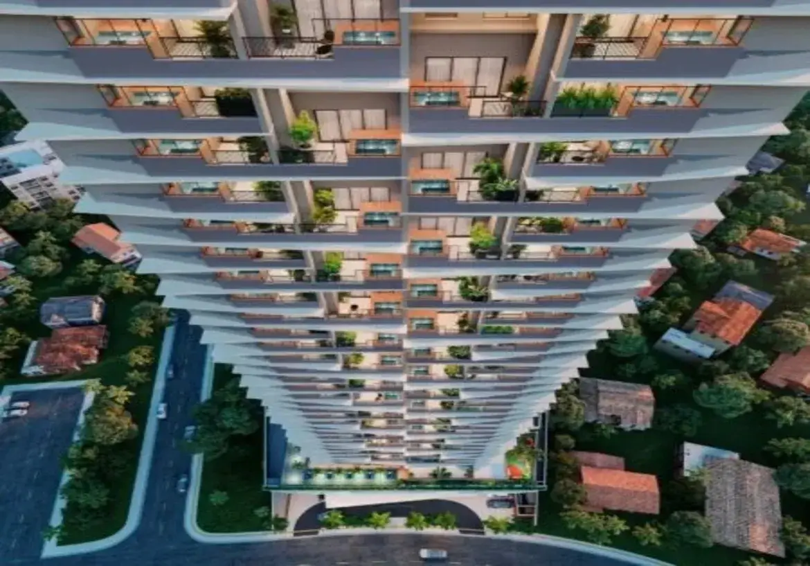 Ok, vamos descrever a imagem detalhadamente: A imagem apresenta uma vista aérea em perspectiva acentuada, olhando de cima para baixo ao longo da fachada de um edifício residencial muito alto e moderno. A perspectiva cria uma sensação de vertigem e destaca a altura imponente do prédio. Detalhes do Imóvel (Edifício): Estrutura e Design: O edifício possui múltiplos andares (um arranha-céu) com uma arquitetura contemporânea. A fachada não é reta, mas sim escalonada ou em degraus, onde cada andar ou conjunto de andares parece ligeiramente deslocado em relação ao inferior/superior, criando um efeito visual dinâmico e texturizado. Cores: A cor predominante da estrutura do edifício é um cinza claro ou branco-acinzentado. As esquadrias das grandes portas de vidro que dão para as varandas e os guarda-corpos parecem ser de um tom escuro, provavelmente preto ou cinza-chumbo. Há também detalhes que parecem ser de madeira clara ou material que imita madeira, visíveis em algumas partes das varandas (possivelmente no piso ou em painéis decorativos/divisórias). Varandas: Cada apartamento possui uma varanda individual. Elas são um elemento central do design. Guarda-corpos: Parecem ser uma combinação de metal escuro com painéis de vidro transparente ou barras verticais finas, garantindo segurança sem obstruir totalmente a vista. Piso: O piso das varandas parece ser claro, talvez cerâmico ou cimentício. Iluminação: A iluminação interna dos apartamentos é visível através das grandes portas de vidro, emitindo uma luz quente e amarelada, sugerindo que a foto foi tirada ao entardecer ou à noite. Algumas varandas também podem ter iluminação própria sutil. Ambiente e Mobiliário (Visível nas Varandas): Móveis: Muitas varandas estão decoradas com móveis de exterior. É possível distinguir pequenas mesas e cadeiras, típicas de conjuntos de bistrô ou áreas de estar compactas. A variedade indica personalização por parte dos moradores. Vegetação: Um detalhe muito relevante é a presença abundante de plantas e flores em vasos em quase todas as varandas. Há vasos de diferentes tamanhos, com plantas de folhagens verdes variadas, algumas mais altas, outras mais baixas, conferindo vida, cor e um aspecto mais natural e acolhedor à fachada predominantemente geométrica e mineral. Ambiente Geral: As luzes acesas e a presença de móveis e plantas sugerem que os apartamentos estão ocupados e as varandas são utilizadas como espaços de convivência e lazer. Entorno: Imediações: O edifício está inserido em uma área urbana, mas com bastante verde. Ao redor da base do prédio, veem-se árvores densas e outras construções mais baixas, principalmente casas com telhados de cerâmica (cor de telha / marrom-avermelhado). Há um contraste nítido entre a escala e modernidade do arranha-céu e as construções vizinhas mais tradicionais. Infraestrutura: Na parte inferior da imagem, são visíveis ruas asfaltadas com alguns carros estacionados e em movimento. Há também calçadas e o acesso à entrada do edifício, que parece ter uma área coberta (porte-cochère) e paisagismo na entrada. Em resumo, a imagem retrata um imponente edifício residencial moderno de cor clara (cinza/branco) com detalhes escuros e em madeira, caracterizado por sua fachada escalonada e varandas individualizadas repletas de plantas. O ambiente combina a verticalidade urbana com a presença significativa de verde, tanto nas varandas quanto no entorno, e a iluminação quente sugere um cenário residencial ao final do dia.