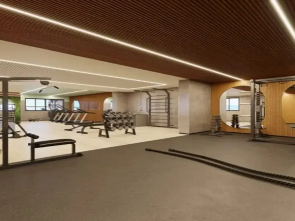 Esta imagem retrata o interior de uma academia de ginástica moderna e bem equipada. O ambiente geral é sofisticado, limpo e funcional, com uma paleta de cores que mistura tons neutros e naturais. Piso: O espaço é dividido por dois tipos de piso. Em primeiro plano e à direita, há um piso emborrachado de cor escura, provavelmente cinza chumbo ou preto, ideal para áreas de peso livre e treinamento funcional. Mais ao fundo e à esquerda, o piso é mais claro, em tons de bege ou creme, possivelmente um vinílico ou porcelanato, cobrindo a área onde ficam as máquinas de cardio e musculação. Paredes: As paredes são predominantemente pintadas em tons claros, como off-white ou bege claro. Destacam-se alguns elementos: À direita, uma parede é revestida com painéis de madeira clara ripada (lambris), onde estão integrados grandes espelhos de formato circular. Isso adiciona um toque natural e amplia o espaço. Uma coluna ou parede apresenta um acabamento texturizado em tom de cimento queimado ou concreto aparente (cinza claro), conferindo um ar industrial moderno. Ao fundo, à esquerda, percebe-se uma parede com revestimento verde vibrante, que parece ser um jardim vertical artificial, trazendo um ponto de cor e natureza para o ambiente. Existem aberturas em formato de arco suave, uma revestida com a mesma madeira clara da parede espelhada, e outras que dão acesso a outras áreas ou janelas. Teto: O teto é um dos destaques do design. Uma grande parte dele é revestida com painéis ripados de madeira escura (tom marrom), conferindo aconchego e sofisticação. A outra seção do teto é rebaixada e pintada de branco. A iluminação principal é feita por longas fitas de LED embutidas que contornam as sancas e as bordas do teto de madeira, proporcionando uma luz brilhante, linear e moderna. Móveis e Equipamentos: A academia está bem equipada com aparelhos modernos, majoritariamente na cor preta e metalizada (cromado ou cinza): Primeiro Plano: Duas grossas cordas navais (battle ropes) pretas estão dispostas no piso escuro. Área Central/Esquerda: Um banco de musculação reto com estrutura preta; um suporte de halteres em formato "A" com vários pares de pesos (pretos e cromados); mais ao fundo, diversas esteiras e possivelmente elípticos. Área Direita/Fundo: Uma estação de musculação multifuncional (possivelmente um crossover ou similar) com estrutura metálica; outro suporte de halteres próximo aos espelhos; na parede com textura de cimento, há um espaldar (barras de parede) metálico. Ambiente e Cores: A combinação do piso escuro funcional, piso claro, paredes neutras, detalhes em madeira clara e escura, o toque de verde e a iluminação LED criam um ambiente ao mesmo tempo energizante e acolhedor. As cores predominantes são os tons terrosos (marrons da madeira), neutros (branco, bege, cinza) e o preto dos equipamentos, com o verde como ponto de destaque. A iluminação abundante e bem distribuída valoriza o design e garante a funcionalidade do espaço.