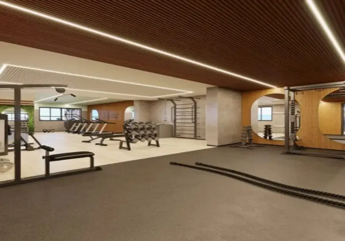 Esta imagem retrata o interior de uma academia de ginástica moderna e bem equipada. O ambiente geral é sofisticado, limpo e funcional, com uma paleta de cores que mistura tons neutros e naturais. Piso: O espaço é dividido por dois tipos de piso. Em primeiro plano e à direita, há um piso emborrachado de cor escura, provavelmente cinza chumbo ou preto, ideal para áreas de peso livre e treinamento funcional. Mais ao fundo e à esquerda, o piso é mais claro, em tons de bege ou creme, possivelmente um vinílico ou porcelanato, cobrindo a área onde ficam as máquinas de cardio e musculação. Paredes: As paredes são predominantemente pintadas em tons claros, como off-white ou bege claro. Destacam-se alguns elementos: À direita, uma parede é revestida com painéis de madeira clara ripada (lambris), onde estão integrados grandes espelhos de formato circular. Isso adiciona um toque natural e amplia o espaço. Uma coluna ou parede apresenta um acabamento texturizado em tom de cimento queimado ou concreto aparente (cinza claro), conferindo um ar industrial moderno. Ao fundo, à esquerda, percebe-se uma parede com revestimento verde vibrante, que parece ser um jardim vertical artificial, trazendo um ponto de cor e natureza para o ambiente. Existem aberturas em formato de arco suave, uma revestida com a mesma madeira clara da parede espelhada, e outras que dão acesso a outras áreas ou janelas. Teto: O teto é um dos destaques do design. Uma grande parte dele é revestida com painéis ripados de madeira escura (tom marrom), conferindo aconchego e sofisticação. A outra seção do teto é rebaixada e pintada de branco. A iluminação principal é feita por longas fitas de LED embutidas que contornam as sancas e as bordas do teto de madeira, proporcionando uma luz brilhante, linear e moderna. Móveis e Equipamentos: A academia está bem equipada com aparelhos modernos, majoritariamente na cor preta e metalizada (cromado ou cinza): Primeiro Plano: Duas grossas cordas navais (battle ropes) pretas estão dispostas no piso escuro. Área Central/Esquerda: Um banco de musculação reto com estrutura preta; um suporte de halteres em formato "A" com vários pares de pesos (pretos e cromados); mais ao fundo, diversas esteiras e possivelmente elípticos. Área Direita/Fundo: Uma estação de musculação multifuncional (possivelmente um crossover ou similar) com estrutura metálica; outro suporte de halteres próximo aos espelhos; na parede com textura de cimento, há um espaldar (barras de parede) metálico. Ambiente e Cores: A combinação do piso escuro funcional, piso claro, paredes neutras, detalhes em madeira clara e escura, o toque de verde e a iluminação LED criam um ambiente ao mesmo tempo energizante e acolhedor. As cores predominantes são os tons terrosos (marrons da madeira), neutros (branco, bege, cinza) e o preto dos equipamentos, com o verde como ponto de destaque. A iluminação abundante e bem distribuída valoriza o design e garante a funcionalidade do espaço.