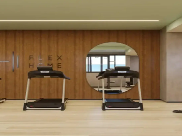 A imagem retrata o interior de uma academia ou espaço fitness de um imóvel, com um design moderno e funcional. Paredes e Revestimentos: A parede principal ao fundo é o destaque visual, revestida com painéis de madeira verticais de tom médio a claro, com veios visíveis. Esse revestimento confere um toque de aconchego e sofisticação ao ambiente. Nesta parede de madeira, está embutido um grande espelho circular que reflete outra parte da academia, mostrando mais equipamentos, uma janela (indicando luz natural e vista externa) e paredes de cor neutra. À esquerda do espelho, na mesma parede de madeira, há a inscrição "FLEX HOME" em letras de forma escuras, dispostas em duas linhas. Mais à esquerda, barras metálicas verticais estão fixadas na parede, sugerindo um espaldar ou equipamento de alongamento/exercício. As paredes laterais e a coluna estrutural visível à direita são pintadas em um tom neutro, provavelmente um cinza muito claro ou bege, criando um contraste suave com a madeira. Piso: O piso é de um material claro, que parece ser laminado de madeira ou vinílico com padrão amadeirado bem claro. Isso contribui para a sensação de limpeza, amplitude e luminosidade do espaço. Teto e Iluminação: O teto é liso e pintado de uma cor clara (possivelmente um branco ou um tom pastel muito suave, talvez um verde bem claro ou bege). Uma linha de luz LED embutida percorre o teto horizontalmente, quase de lado a lado, proporcionando uma iluminação linear, difusa e moderna. Móveis e Equipamentos: Em primeiro plano, duas esteiras ergométricas estão posicionadas. Elas possuem estrutura predominantemente preta (plataforma, console) com detalhes e hastes laterais em metal prateado/cinza claro. Parecem ser equipamentos modernos e de boa qualidade. O reflexo no espelho mostra pelo menos mais uma esteira e possivelmente outros aparelhos de musculação ou cardio, além de um tapete de yoga ou colchonete azul no chão. Ambiente e Cores: O ambiente geral é clean, moderno e bem iluminado. Transmite uma sensação de espaço funcional e agradável para a prática de exercícios. A paleta de cores é predominantemente neutra e natural: tons de madeira (médio na parede, claro no piso), cinza/bege claro nas outras paredes, branco/claro no teto, preto e prata nos equipamentos. A combinação cria um espaço equilibrado e convidativo. Em resumo, trata-se de uma academia bem equipada e com design contemporâneo, utilizando madeira para aquecer o ambiente, cores neutras para ampliar e espelhos/iluminação para funcionalidade e estética.