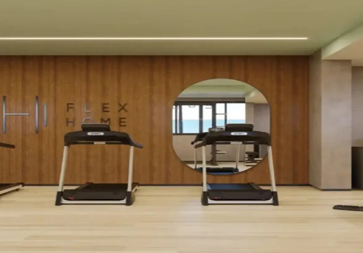 A imagem retrata o interior de uma academia ou espaço fitness de um imóvel, com um design moderno e funcional. Paredes e Revestimentos: A parede principal ao fundo é o destaque visual, revestida com painéis de madeira verticais de tom médio a claro, com veios visíveis. Esse revestimento confere um toque de aconchego e sofisticação ao ambiente. Nesta parede de madeira, está embutido um grande espelho circular que reflete outra parte da academia, mostrando mais equipamentos, uma janela (indicando luz natural e vista externa) e paredes de cor neutra. À esquerda do espelho, na mesma parede de madeira, há a inscrição "FLEX HOME" em letras de forma escuras, dispostas em duas linhas. Mais à esquerda, barras metálicas verticais estão fixadas na parede, sugerindo um espaldar ou equipamento de alongamento/exercício. As paredes laterais e a coluna estrutural visível à direita são pintadas em um tom neutro, provavelmente um cinza muito claro ou bege, criando um contraste suave com a madeira. Piso: O piso é de um material claro, que parece ser laminado de madeira ou vinílico com padrão amadeirado bem claro. Isso contribui para a sensação de limpeza, amplitude e luminosidade do espaço. Teto e Iluminação: O teto é liso e pintado de uma cor clara (possivelmente um branco ou um tom pastel muito suave, talvez um verde bem claro ou bege). Uma linha de luz LED embutida percorre o teto horizontalmente, quase de lado a lado, proporcionando uma iluminação linear, difusa e moderna. Móveis e Equipamentos: Em primeiro plano, duas esteiras ergométricas estão posicionadas. Elas possuem estrutura predominantemente preta (plataforma, console) com detalhes e hastes laterais em metal prateado/cinza claro. Parecem ser equipamentos modernos e de boa qualidade. O reflexo no espelho mostra pelo menos mais uma esteira e possivelmente outros aparelhos de musculação ou cardio, além de um tapete de yoga ou colchonete azul no chão. Ambiente e Cores: O ambiente geral é clean, moderno e bem iluminado. Transmite uma sensação de espaço funcional e agradável para a prática de exercícios. A paleta de cores é predominantemente neutra e natural: tons de madeira (médio na parede, claro no piso), cinza/bege claro nas outras paredes, branco/claro no teto, preto e prata nos equipamentos. A combinação cria um espaço equilibrado e convidativo. Em resumo, trata-se de uma academia bem equipada e com design contemporâneo, utilizando madeira para aquecer o ambiente, cores neutras para ampliar e espelhos/iluminação para funcionalidade e estética.