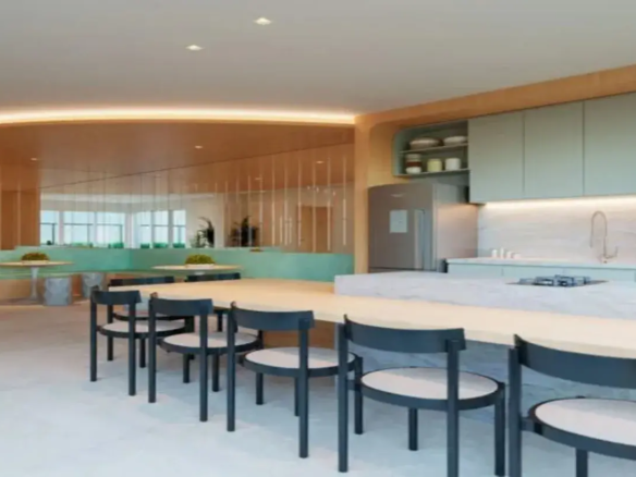 A imagem retrata um ambiente interno moderno e funcional, provavelmente uma copa, cozinha gourmet ou área de convivência de um prédio residencial, co-living ou escritório. O espaço é bem iluminado e apresenta um design clean com uma paleta de cores suaves e materiais contemporâneos. Principais Elementos e Detalhes: Mobiliário Principal (Primeiro Plano): Uma mesa longa e retangular com tampo em madeira clara (ou laminado que imita madeira) ocupa o centro do primeiro plano. Serve como uma grande mesa de jantar ou bancada de trabalho/refeições comunitária. Ao redor da mesa, há várias cadeiras de design simples e moderno. Elas possuem estrutura preta (provavelmente metal ou madeira pintada) e assentos redondos estofados em um tecido de cor clara, como bege ou off-white. Área da Cozinha (Lado Direito): Uma bancada extensa em material que imita mármore branco ou quartzo claro, com veios suaves acinzentados. Esta bancada serve como área de preparo e também se estende formando um balcão voltado para a mesa principal. Embutido na bancada, há um cooktop (fogão de mesa). Mais ao fundo, na mesma bancada, percebe-se uma pia com uma torneira de design moderno, possivelmente em acabamento metálico (talvez cobre ou dourado fosco). O frontão (backsplash) da pia segue o mesmo material da bancada, criando continuidade visual. Armários superiores com portas lisas em um tom suave de verde-menta ou azul-petróleo claro. Ao lado dos armários, acima da geladeira, há um nicho aberto com prateleiras, feito em madeira clara, combinando com a mesa e outros detalhes. Contém alguns potes ou itens decorativos. Uma geladeira em aço inoxidável (inox) está posicionada sob o nicho. Iluminação sob os armários destaca a bancada e o frontão. Área de Estar Secundária (Fundo/Lado Esquerdo): Uma parede revestida com painéis espelhados verticais ou um grande espelho segmentado. Isso amplia visualmente o espaço e reflete a área oposta. No reflexo (e possivelmente na área real atrás da mesa principal), há uma área de estar com bancos/booths estofados no mesmo tom de verde-menta/azul-petróleo claro dos armários da cozinha. A base desses bancos parece ser de madeira clara. Mesas redondas pequenas com tampo de madeira clara e base central estão dispostas junto aos bancos. Alguns bancos/pufes cilíndricos de cor cinza texturizada complementam essa área de estar. Pequenos vasos com plantas verdes decoram as mesas menores. No reflexo do espelho, é possível ver janelas amplas ao fundo, indicando entrada de luz natural. Piso e Teto: O piso é revestido com ladrilhos ou porcelanato de grande formato em tom claro, provavelmente cinza-claro ou off-white, conferindo um aspecto limpo e neutro. O teto é branco e liso, com iluminação embutida (spots de luz quadrados) e uma sanca de luz indireta contornando uma estrutura rebaixada em madeira clara acima da parede espelhada, proporcionando uma iluminação ambiente suave e quente. Cores e Ambiente: A paleta de cores é predominantemente neutra e suave, combinando tons de madeira clara, branco, cinza-claro, com toques de verde-menta/azul-petróleo claro nos armários e estofados, e preto nas estruturas das cadeiras para contraste. O uso de espelhos e a iluminação bem planejada (natural e artificial) criam um ambiente claro, arejado, moderno e acolhedor. O design é funcional e pensado para o convívio social ou refeições informais.