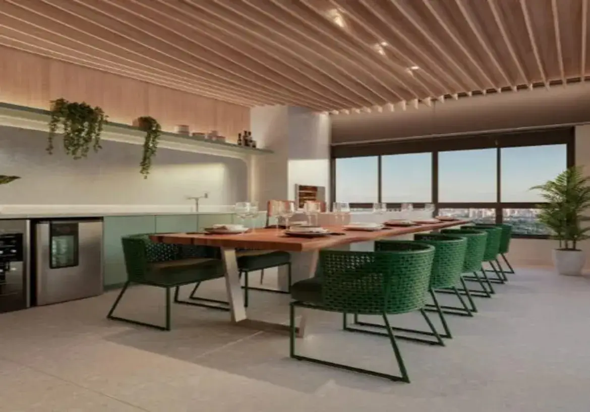 A imagem retrata uma espaçosa e moderna área de jantar, possivelmente integrada a uma varanda gourmet ou área de convivência em um apartamento de alto padrão. Ambiente e Cores: O ambiente geral é sofisticado e contemporâneo, com um toque natural. A paleta de cores principal combina tons neutros e terrosos com um verde vibrante. O piso é de um material claro, possivelmente porcelanato em tom de bege ou cinza muito claro, o que contribui para a sensação de amplitude e luminosidade. As paredes são predominantemente brancas ou off-white. O teto é um destaque, revestido com ripas de madeira clara (ripado de madeira), conferindo aconchego e um design diferenciado, com iluminação embutida entre as ripas. Grandes janelas de vidro com esquadrias escuras (pretas ou grafite) ocupam uma parede inteira, oferecendo uma vista panorâmica da cidade e permitindo a entrada de bastante luz natural. Há persianas rolô recolhidas na parte superior das janelas. Móveis: Mesa de Jantar: O ponto central é uma longa mesa de jantar retangular. O tampo é robusto, feito de madeira maciça com acabamento natural, possivelmente com bordas orgânicas (live edge), exibindo os veios da madeira. A base da mesa é feita de metal, com pés em design moderno e angular, em cor clara (prata ou aço escovado). Cadeiras: Ao redor da mesa, há várias cadeiras de design moderno. Elas possuem uma estrutura fina de metal em cor escura (preta ou grafite) e o assento e encosto são revestidos em um material com trama ou textura, numa cor verde-escuro ou verde-musgo vibrante, que contrasta elegantemente com a madeira da mesa e os tons neutros do ambiente. São visíveis pelo menos 8 cadeiras. Bancada/Armários: Ao fundo, estendendo-se por uma parede, há uma bancada longa e baixa com armários inferiores em um tom suave de verde-claro (verde-água ou menta). Acima da bancada, há uma prateleira comprida na mesma cor dos armários inferiores, decorada com algumas plantas pendentes e objetos. Abaixo da bancada, à esquerda, notam-se eletrodomésticos embutidos com acabamento em aço inoxidável, parecendo ser uma adega climatizada ou frigobar. Há também uma torneira visível sobre a bancada, indicando a presença de uma pia. Detalhes e Decoração: Iluminação: Além da luz natural das janelas, a iluminação artificial vem dos spots embutidos no teto ripado e de uma fita de LED sob a prateleira, que ilumina a área da bancada. A iluminação geral parece ser quente e convidativa. Plantas: Elementos verdes naturais estão presentes: duas plantas pendentes na prateleira e uma planta maior em um vaso branco no canto direito, próximo à janela, adicionando vida e frescor ao espaço. Mesa Posta: A mesa está parcialmente posta com pratos brancos, taças de vinho e copos de água, sugerindo que o espaço está pronto para receber convidados ou para uso. Em resumo, o imóvel apresenta um design de interiores moderno, que equilibra materiais naturais como a madeira com elementos contemporâneos como o metal e o vidro. A combinação de cores neutras com o verde vibrante das cadeiras e o verde suave da bancada cria um espaço visualmente interessante, funcional e acolhedor, ideal para refeições e convívio social.