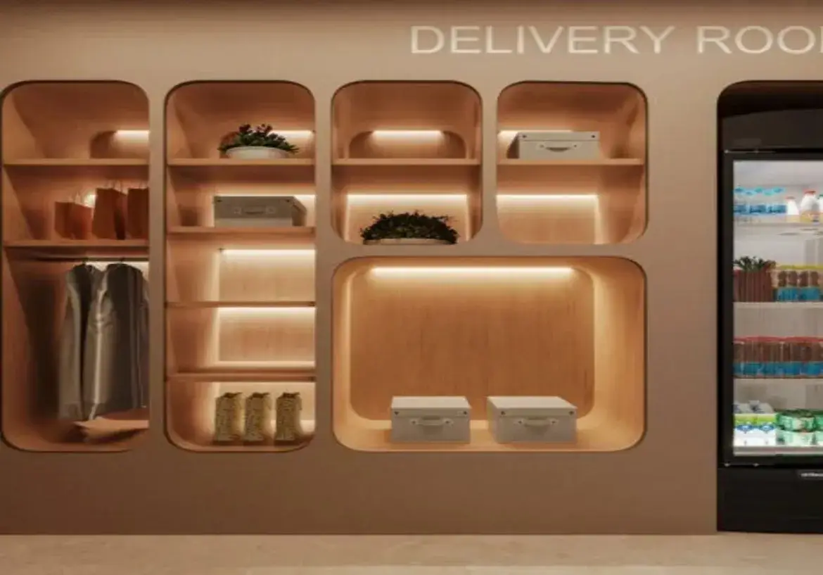 A imagem apresenta um espaço moderno e organizado identificado como "DELIVERY ROOM" (Sala de Entregas), provavelmente localizado na área comum de um edifício residencial ou comercial. Estrutura Principal e Cores: A estrutura principal é uma grande unidade de parede embutida, que combina nichos de armazenamento abertos e um refrigerador. A cor predominante da estrutura externa é um bege ou taupe claro, com um acabamento fosco. Os interiores dos nichos são revestidos com um material que imita madeira clara, criando um contraste suave e acolhedor com a cor externa. O piso visível na parte inferior é de um material claro, possivelmente porcelanato ou concreto polido, em tom neutro. Móveis e Armazenamento: O móvel central é a estante embutida, caracterizada por múltiplos nichos de tamanhos variados, todos com cantos arredondados, conferindo um design suave e contemporâneo. Nichos: Existem vários nichos: Um nicho vertical alto à esquerda, dividido em duas partes: a superior contém sacolas de papel marrons e a inferior possui um varão com algumas peças de roupa penduradas (aparentemente camisas ou casacos em tons neutros/cinza). Uma coluna central com várias prateleiras horizontais. Contém: um vaso branco com uma planta na prateleira superior, uma caixa de armazenamento cinza claro com alça na prateleira abaixo, e na prateleira inferior, o que parecem ser três objetos decorativos ou talvez botas texturizadas em tom claro. Nichos menores, quadrados ou retangulares, localizados na parte superior e central direita. Um deles contém outra planta em vaso branco, e outro contém uma caixa de armazenamento cinza claro idêntica à da coluna central. Um nicho horizontal grande na parte inferior direita, contendo duas caixas de armazenamento cinza claro com alças, lado a lado. Refrigerador: À direita da unidade de nichos, há um refrigerador alto com porta de vidro e moldura preta. Ele parece estar bem abastecido com bebidas (garrafas, caixas de leite/sumo, latas) organizadas nas prateleiras internas. A iluminação interna do refrigerador é mais fria (branca) em contraste com a iluminação quente dos nichos. Iluminação e Ambiente: A iluminação é um destaque no design. A maioria das prateleiras horizontais dentro dos nichos possui fitas de LED embutidas na parte inferior, projetando uma luz quente e indireta para baixo. Isso não só ilumina os objetos armazenados, mas também cria um ambiente acolhedor, sofisticado e convidativo. A iluminação geral do espaço parece suave. Decoração e Detalhes: Além dos itens funcionais (caixas, roupas, bebidas), há elementos decorativos como as plantas em vasos brancos, que adicionam um toque de vida e cor ao ambiente predominantemente neutro. As caixas de armazenamento cinza claro com alças contribuem para a sensação de ordem e padronização. A inscrição "DELIVERY ROOM" em letras brancas, simples e maiúsculas, na parte superior da estrutura, identifica claramente a função do espaço. Impressão Geral: O ambiente transmite uma sensação de modernidade, funcionalidade e organização. A paleta de cores neutras e quentes (bege, madeira clara, cinza claro) combinada com a iluminação indireta e o design de linhas suaves (cantos arredondados) cria um espaço acolhedor e esteticamente agradável, pensado para facilitar o recebimento e armazenamento temporário de entregas de forma prática e elegante.