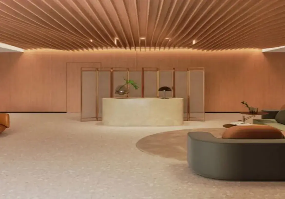 A imagem retrata um ambiente interior amplo e sofisticado, provavelmente um lobby de hotel, recepção de um edifício comercial de luxo ou uma área de espera de alto padrão. A atmosfera geral é calma, elegante e moderna, com um forte foco em design e materiais de qualidade. Detalhes Relevantes: Teto: O teto é um dos elementos mais marcantes. É composto por ripas de madeira paralelas, em um tom claro e quente (semelhante a carvalho ou pinus claro), que seguem um design ondulado e orgânico. Esta forma curva e fluida cria um efeito visual dinâmico e acolhedor. A iluminação está integrada ao teto, com pontos de luz embutidos (spots) discretamente posicionados entre as ripas e uma iluminação indireta (sanca de luz) na junção do teto com as paredes, proporcionando uma luz suave e difusa que realça a textura da madeira e as curvas. Paredes: As paredes visíveis são revestidas com painéis lisos, possivelmente de madeira ou um material similar, em um tom suave de terracota ou rosa queimado. Essa cor quente complementa o tom da madeira do teto e contribui para a sensação acolhedora do espaço. Piso: O piso é claro, parecendo ser de granilite ou um material composto similar (terrazzo), com uma base bege ou branco-acinzentada salpicada de pequenos agregados em tons mais escuros e neutros. Confere um ar moderno e é fácil de manter, além de refletir suavemente a luz. Mobiliário e Layout: Recepção/Balcão: Ao fundo, centralizado, há um balcão de recepção curvo, com linhas suaves. Ele parece ser feito de mármore ou um material similar em tom creme ou bege muito claro, com uma superfície polida. Sobre o balcão, há um vaso de vidro arredondado com um arranjo simples de folhas verdes e um candeeiro de mesa moderno com cúpula escura e base metálica. Divisória: Atrás do balcão, uma divisória decorativa (biombo) composta por vários painéis verticais com molduras metálicas finas (possivelmente em tom de latão ou cobre rosado) e um preenchimento translúcido ou em tecido claro (bege/off-white) ajuda a definir o espaço sem bloqueá-lo completamente. Sofás: No primeiro plano e à direita, há uma área de estar. Um sofá curvo, com design contemporâneo e estofado em um tecido de cor verde-acinzentado escuro ou musgo, ocupa parte da cena. Sobre ele, uma almofada em tom terroso (laranja queimado ou terracota) adiciona um ponto de cor. Mais ao fundo, outro sofá de design similar é parcialmente visível. Sob o sofá do primeiro plano, um tapete redondo em tom neutro (bege claro) ajuda a delimitar a área de estar. Poltrona: À esquerda, parcialmente visível, há o que parece ser uma poltrona de couro em tom caramelo ou conhaque, também com design moderno. Mesa de Centro: Uma pequena mesa de centro baixa com tampo escuro e pés finos está posicionada perto do sofá do fundo. Cores e Ambiente: A paleta de cores é predominantemente quente e terrosa, com os tons de madeira clara, terracota/rosa queimado, creme, bege, e acentos em verde musgo e laranja queimado. Os detalhes metálicos da divisória adicionam um toque de sofisticação. A iluminação indireta e quente, combinada com as formas curvas (teto, balcão, sofás) e os materiais naturais (madeira, pedra, couro), cria um ambiente muito acolhedor, sofisticado, tranquilo e contemporâneo.