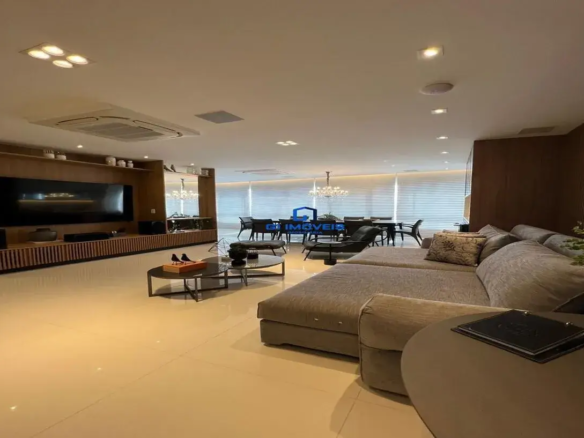 Esta imagem retrata um ambiente interno amplo, moderno e sofisticado, que integra sala de estar e sala de jantar em um conceito aberto. Cores e Materiais: A paleta de cores é predominantemente neutra e elegante, com tons de bege, cinza e marrom. Piso: O chão é revestido com grandes placas de porcelanato em tom bege claro ou creme, com acabamento polido que reflete a luz, contribuindo para a sensação de amplitude. Paredes e Teto: O teto é branco ou off-white, com iluminação embutida. Uma das paredes (à direita e ao fundo, atrás do sofá) é revestida com um painel de madeira ripada em tom marrom escuro, conferindo textura e aconchego ao ambiente. Móveis: Predominam tons de cinza e marrom. O sofá é cinza claro, a madeira dos painéis e do rack da TV é marrom, e há detalhes em preto nas mesas de centro e cadeiras da sala de jantar. Ambiente e Iluminação: O ambiente é luminoso e bem iluminado, tanto por luz natural (proveniente de grandes janelas ao fundo, cobertas por persianas horizontais claras) quanto artificial. Iluminação Artificial: O teto possui diversos pontos de luz embutidos (spots), incluindo um conjunto de quatro spots quadrados próximos à área da TV e outros distribuídos pelo espaço. Um lustre de cristal imponente e clássico pende sobre a mesa de jantar, adicionando um toque de requinte. Climatização: Um grande aparelho de ar condicionado central do tipo cassete está embutido no teto. Atmosfera: A atmosfera geral é de sofisticação, conforto e modernidade, com um design de interiores clean e bem planejado. Móveis e Decoração: Sala de Estar: Sofá: Um grande sofá modular em "L" de tecido cinza claro domina o primeiro plano. Possui almofadas grandes e algumas decorativas, uma delas com estampa sutil em tons de bege e marrom. Mesa de Apoio Lateral: Ao lado do sofá, uma mesa de apoio redonda em tom cinza escuro ou taupe fosco, com um objeto escuro (possivelmente um livro ou caixa decorativa) sobre ela. Mesas de Centro: Duas mesas de centro ovais/arredondadas, sendo uma ligeiramente menor e mais baixa que a outra (estilo ninho). Possuem estrutura metálica escura (preta) e tampos que parecem ser de vidro escuro ou material similar. Sobre a mesa maior, há um pequeno arranjo com duas estatuetas de pássaros pretos com detalhes em laranja/marrom. Painel de TV e Rack: À esquerda, um grande painel de madeira que vai do chão ao teto acomoda uma TV de tela plana. Abaixo da TV, um rack baixo e comprido, também em madeira com detalhes ripados, percorre a extensão do painel. Prateleiras embutidas no painel exibem objetos decorativos, como vasos brancos e outras estatuetas de pássaros. Espelho: Ao lado do painel da TV, um grande espelho vertical reflete parte da sala de jantar, ampliando visualmente o espaço. Sala de Jantar (ao fundo): Mesa de Jantar: Uma mesa de jantar retangular grande, com tampo escuro (possivelmente preto ou madeira escura). Cadeiras: Diversas cadeiras de design moderno com estrutura escura e assento/encosto em tom claro. Poltrona/Chaise: Entre a sala de estar e a de jantar, vislumbra-se uma poltrona de design ou chaise longue preta com estrutura metálica. O imóvel demonstra um projeto de interiores contemporâneo, que valoriza espaços integrados, iluminação abundante e uma paleta de cores neutras com texturas ricas, resultando em um ambiente elegante e convidativo.